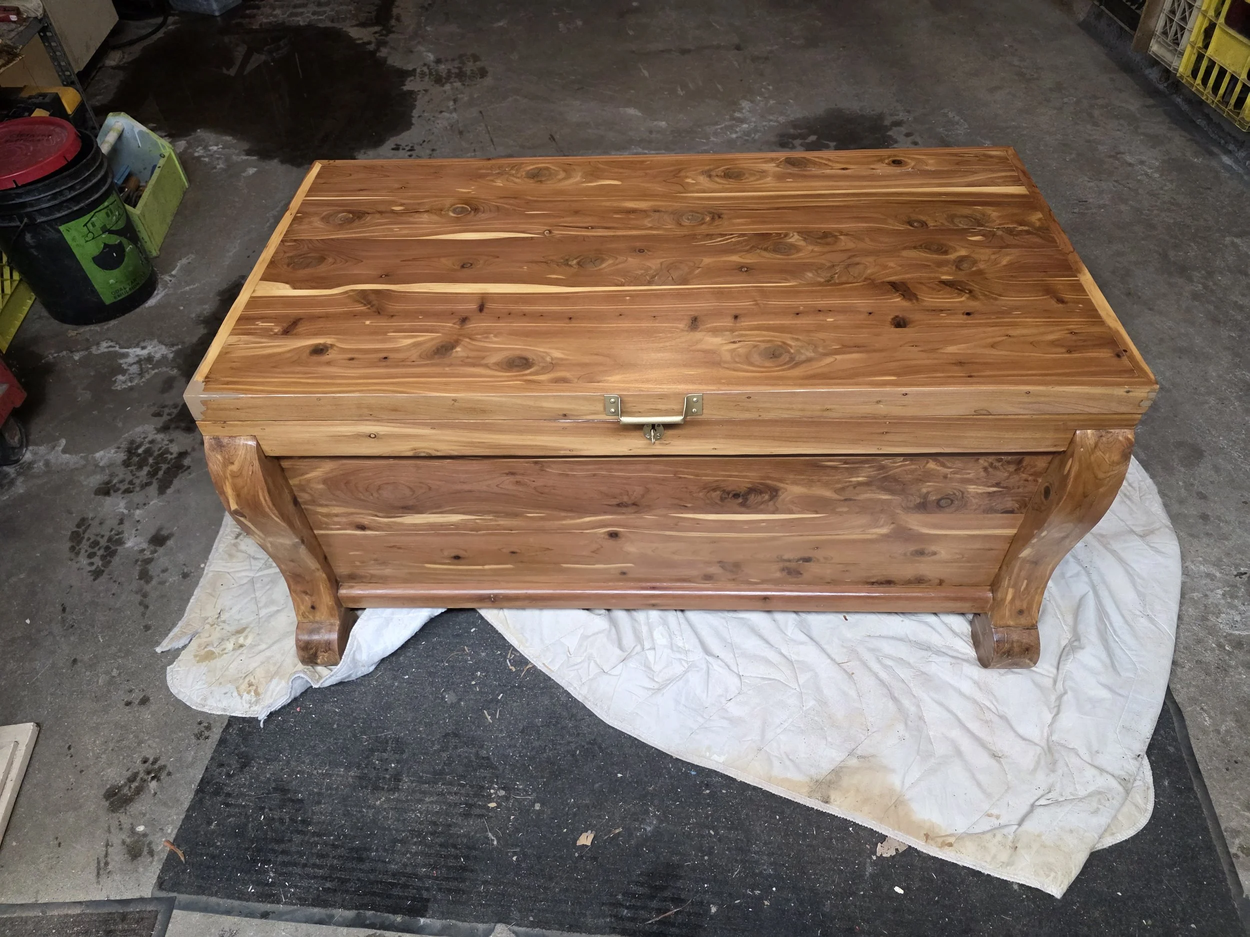 A wooden chest with a latch on the front, placed on a white cloth on a concrete floor in a workshop.