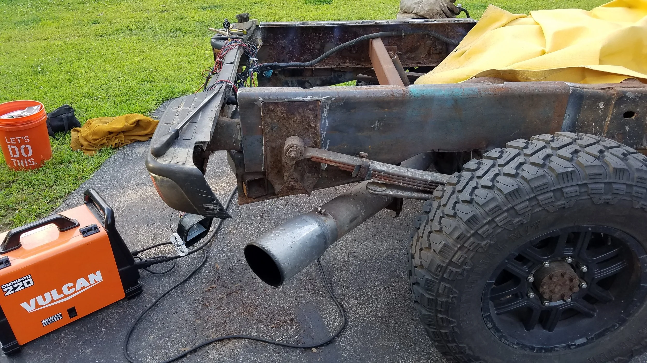 A vehicle's chassis with large off-road tires, a muffler, and various tools and equipment nearby, including an orange welding machine and a bucket on grass.
