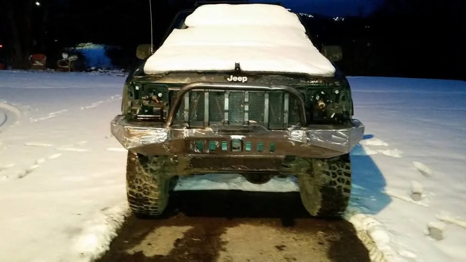 Front view of a black Jeep with a snow-covered roof, parked on a snowy ground at night, with some damage and tape on the front bumper.