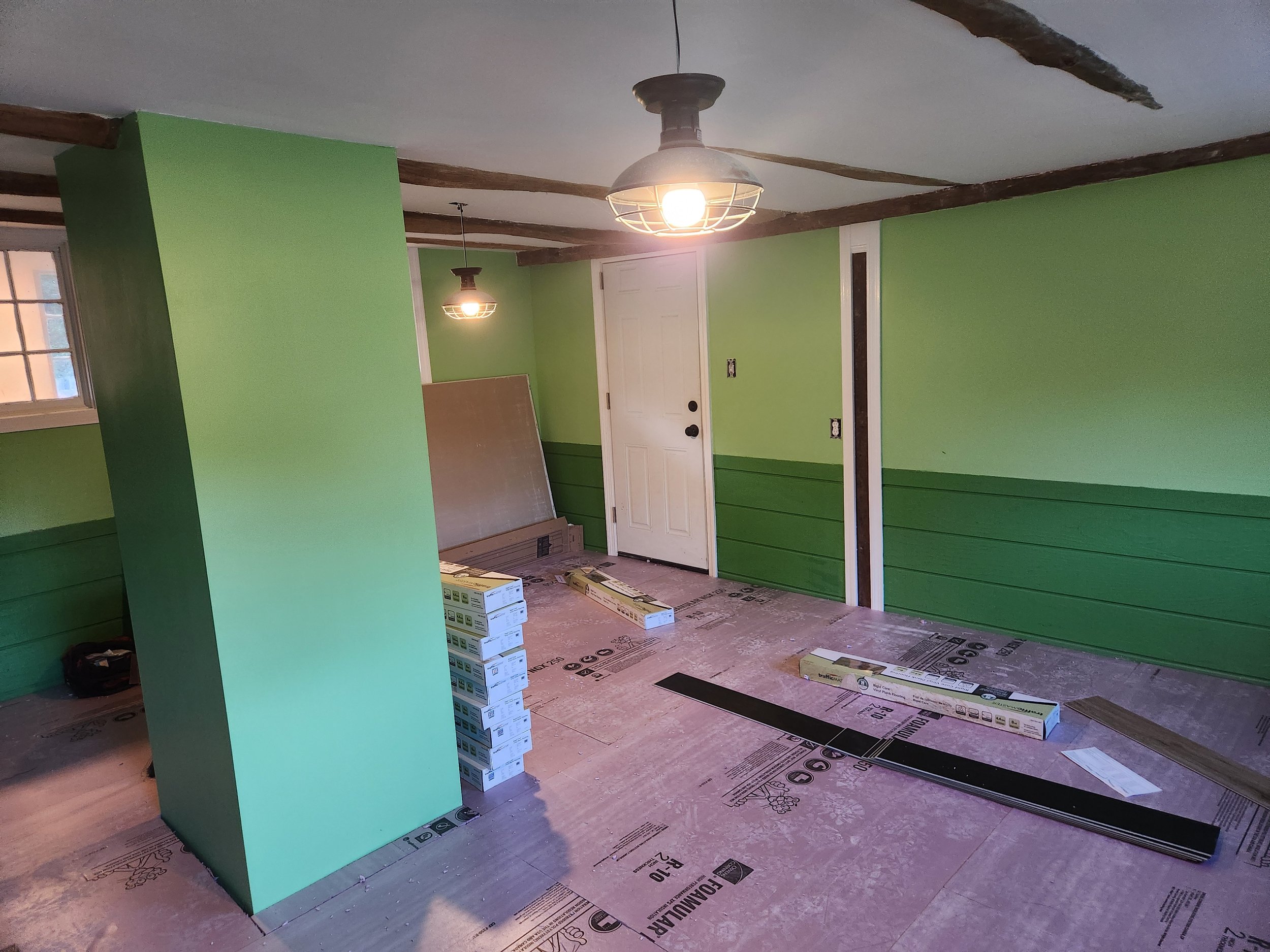 An interior room under renovation with light green walls, wooden ceiling beams, and a ceiling fan. There are construction materials and boxes on the floor, and the floor is covered with protective sheeting.