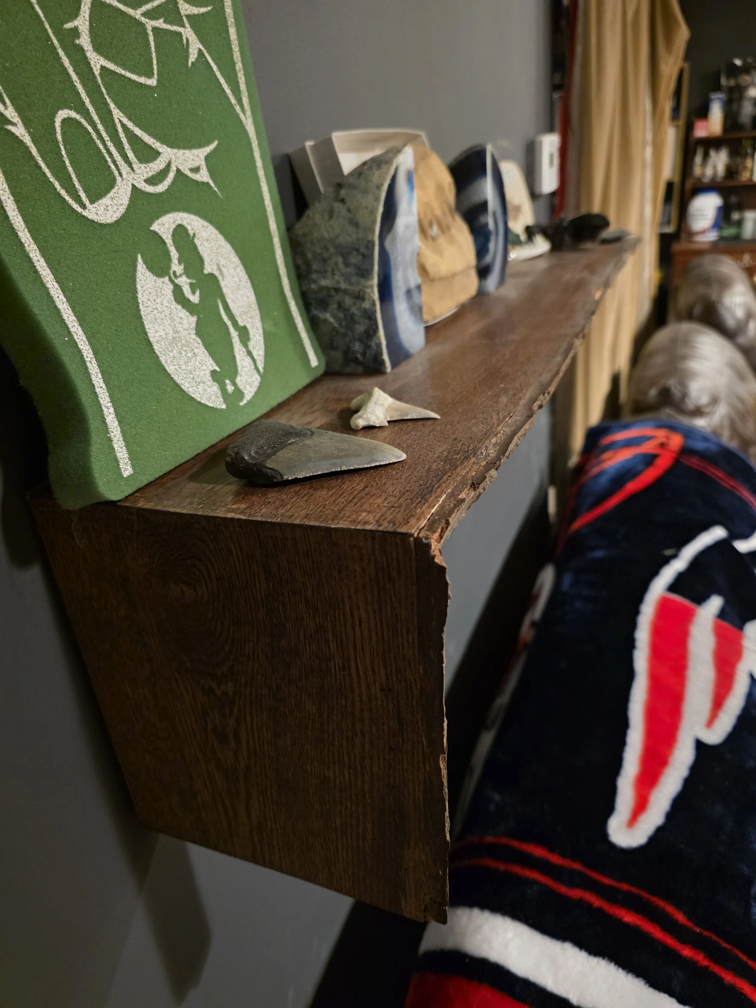 Close-up of a wooden shelf with various stones and artwork, including a green piece with a white silhouette, against a dark gray wall in a room.