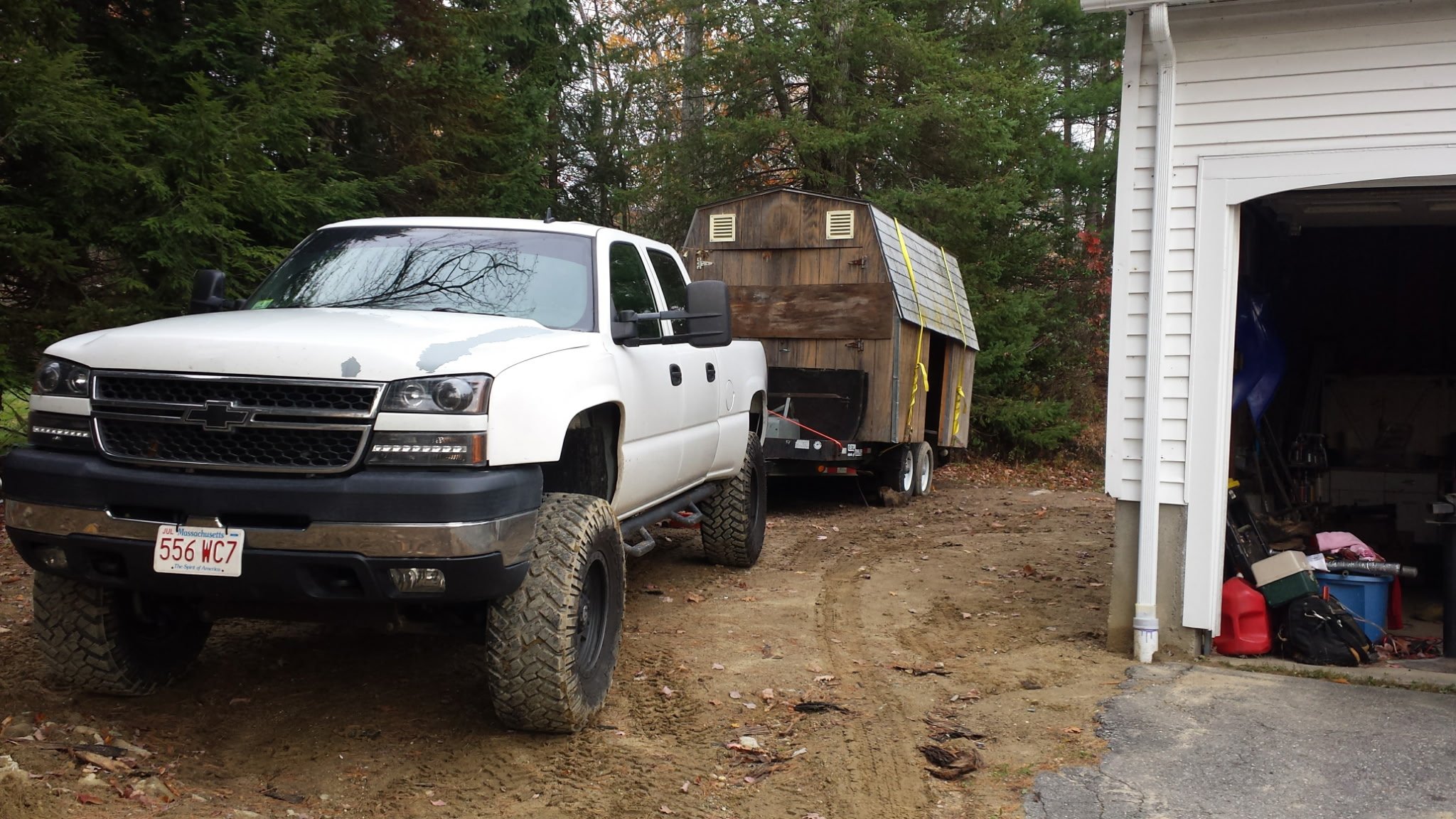 White pickup truck parked outside a garage with a wooden trailer attached, surrounded by trees and dirt ground.