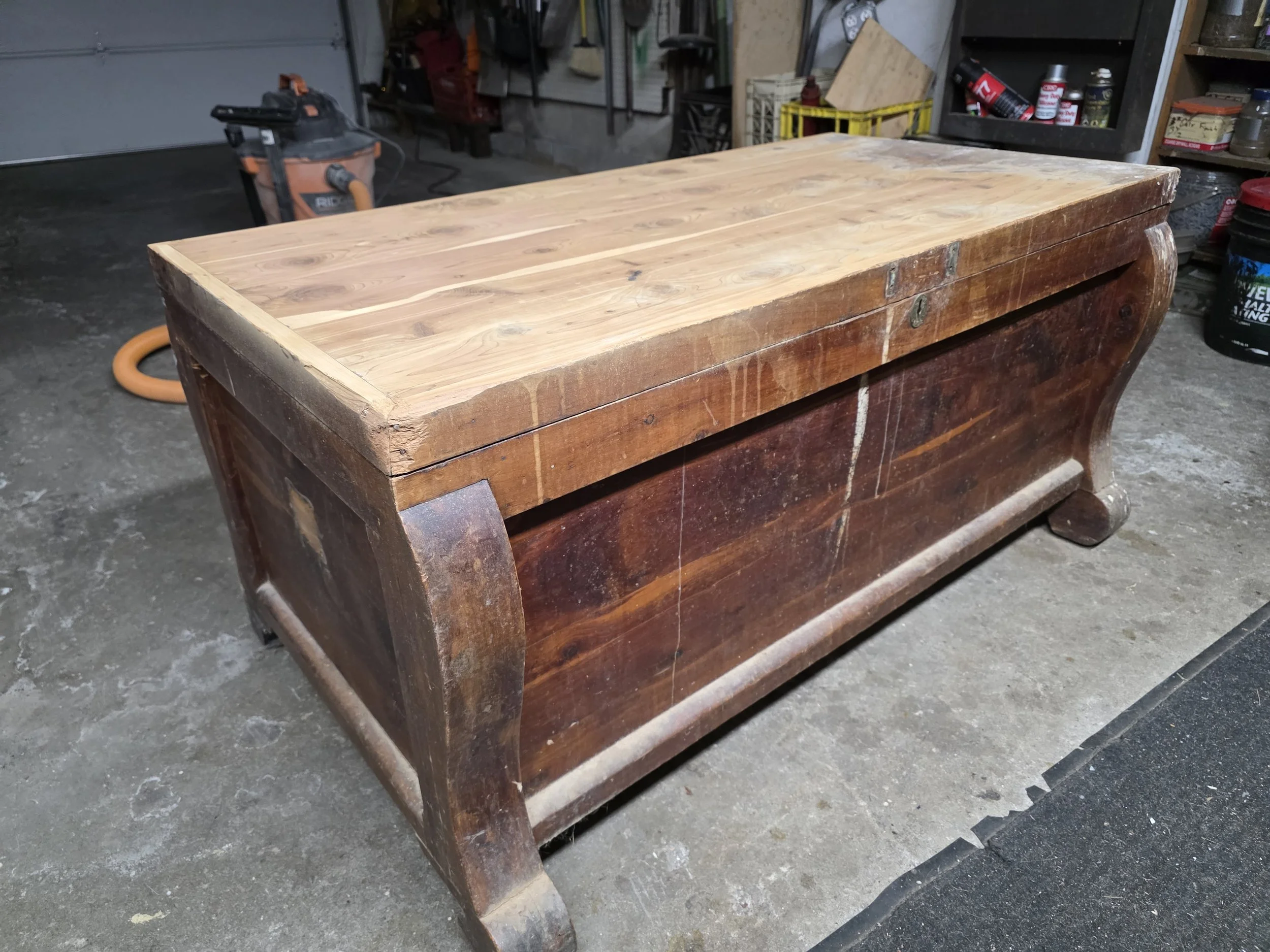 A wooden chest with a newer, lighter wood top and darker, aged wood sides, sitting on a concrete floor in a workshop.