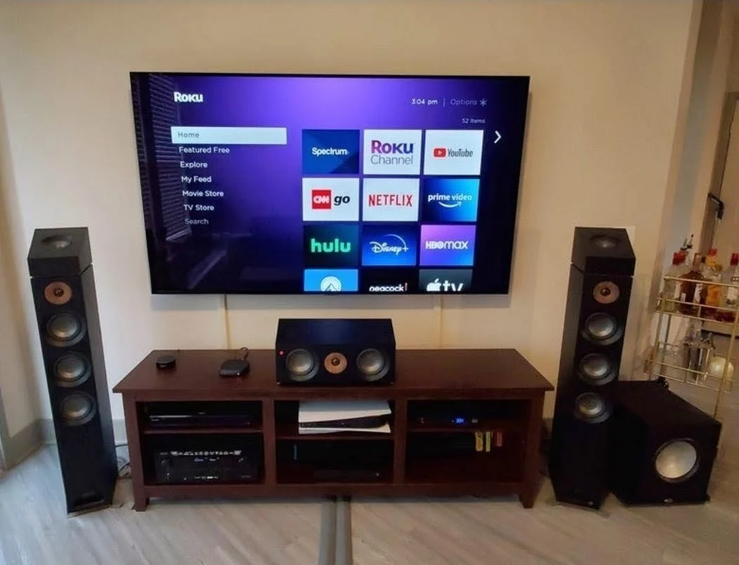 Living room with a flat-screen TV displaying streaming app icons, two tall floor speakers, a center speaker, a subwoofer, and a wooden entertainment stand holding electronic devices.