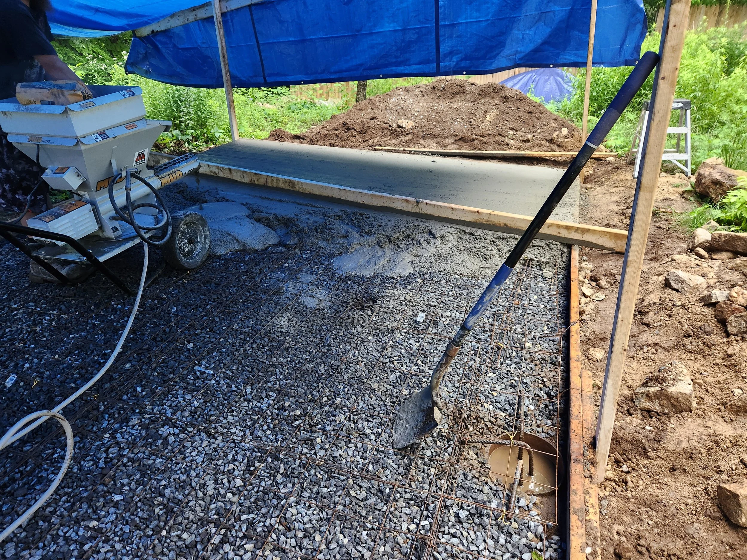 Concrete being poured onto a gravel and rebar foundation under a blue tarp roof, with a shovel and a step ladder visible in the background.