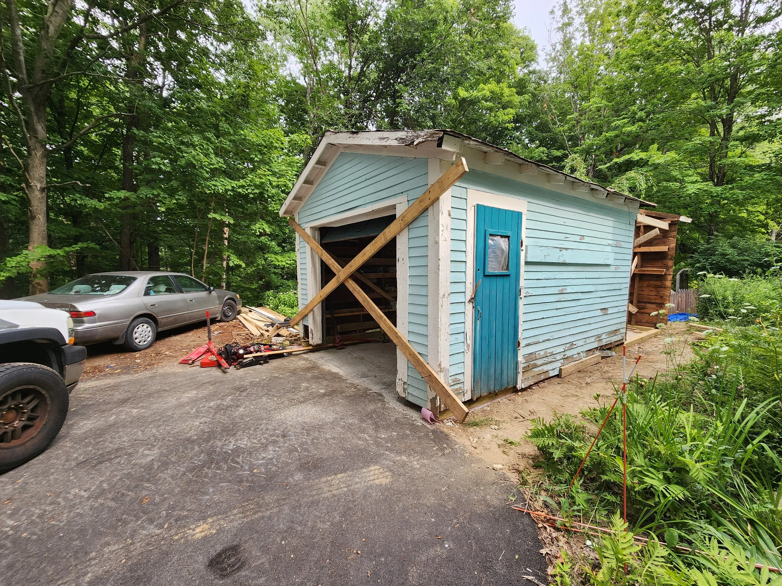 A small blue shed under construction with wooden braces on its front and side, situated by a paved area with two parked cars and surrounded by green trees and plants.