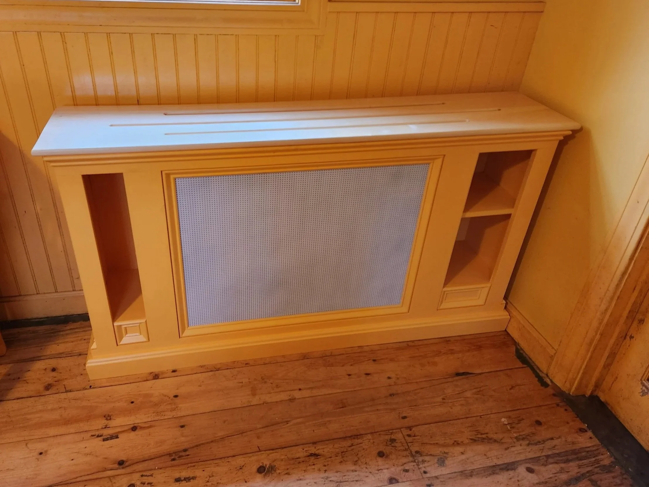 A wooden entertainment center with open shelves and a large mesh-covered opening, placed against a wood-paneled wall on a wooden floor.