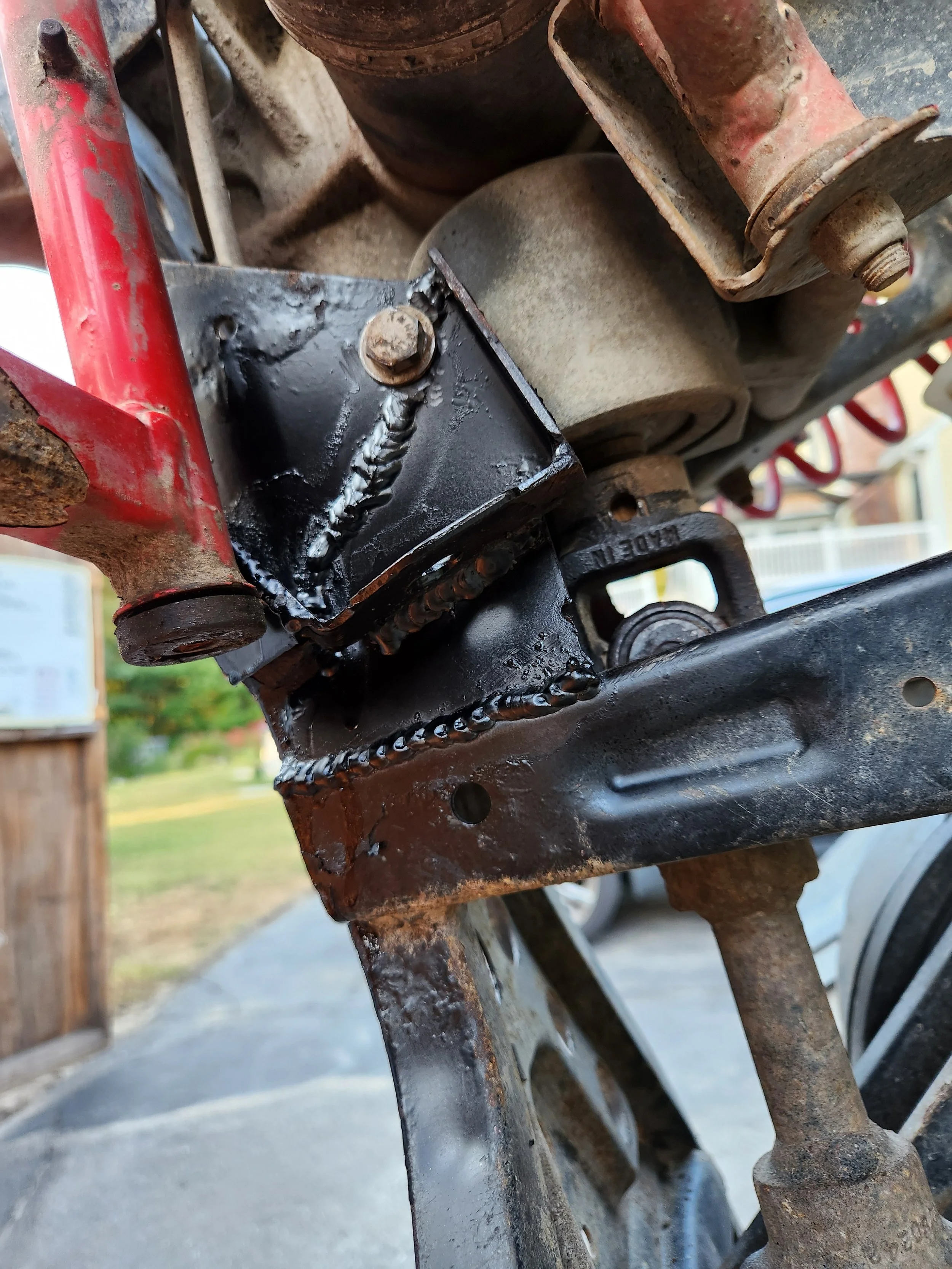 Close-up of a welded metal bracket mounted on a vehicle frame, showing new welding work on the black-painted bracket with some rusted parts nearby.