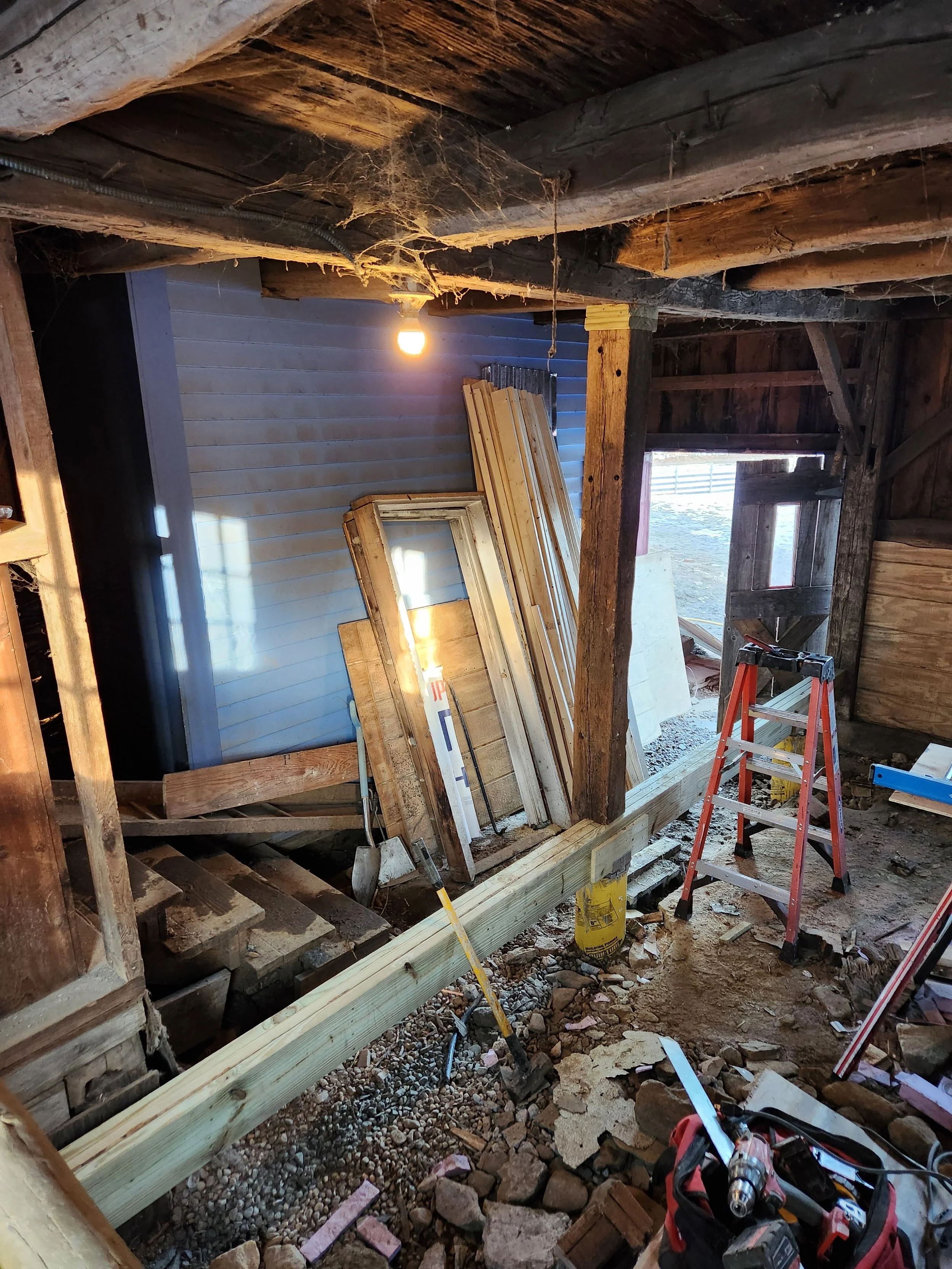 Inside a building under renovation with exposed wooden beams and framing. Construction tools, a step ladder, and stacks of wooden planks are visible, with a single light bulb hanging from the ceiling.