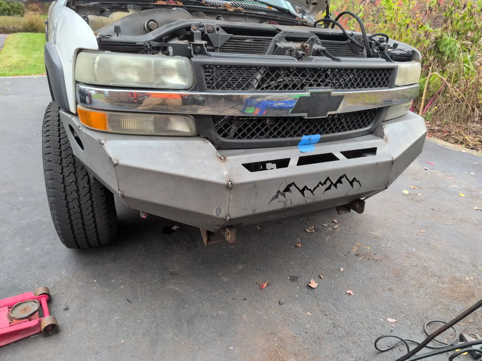Front of a vehicle with a partially disassembled front bumper, exposing the grill and headlights. The bumper is made of metal with mountain range graphics at the bottom. There is a blue paint marker and some tools on the ground nearby.
