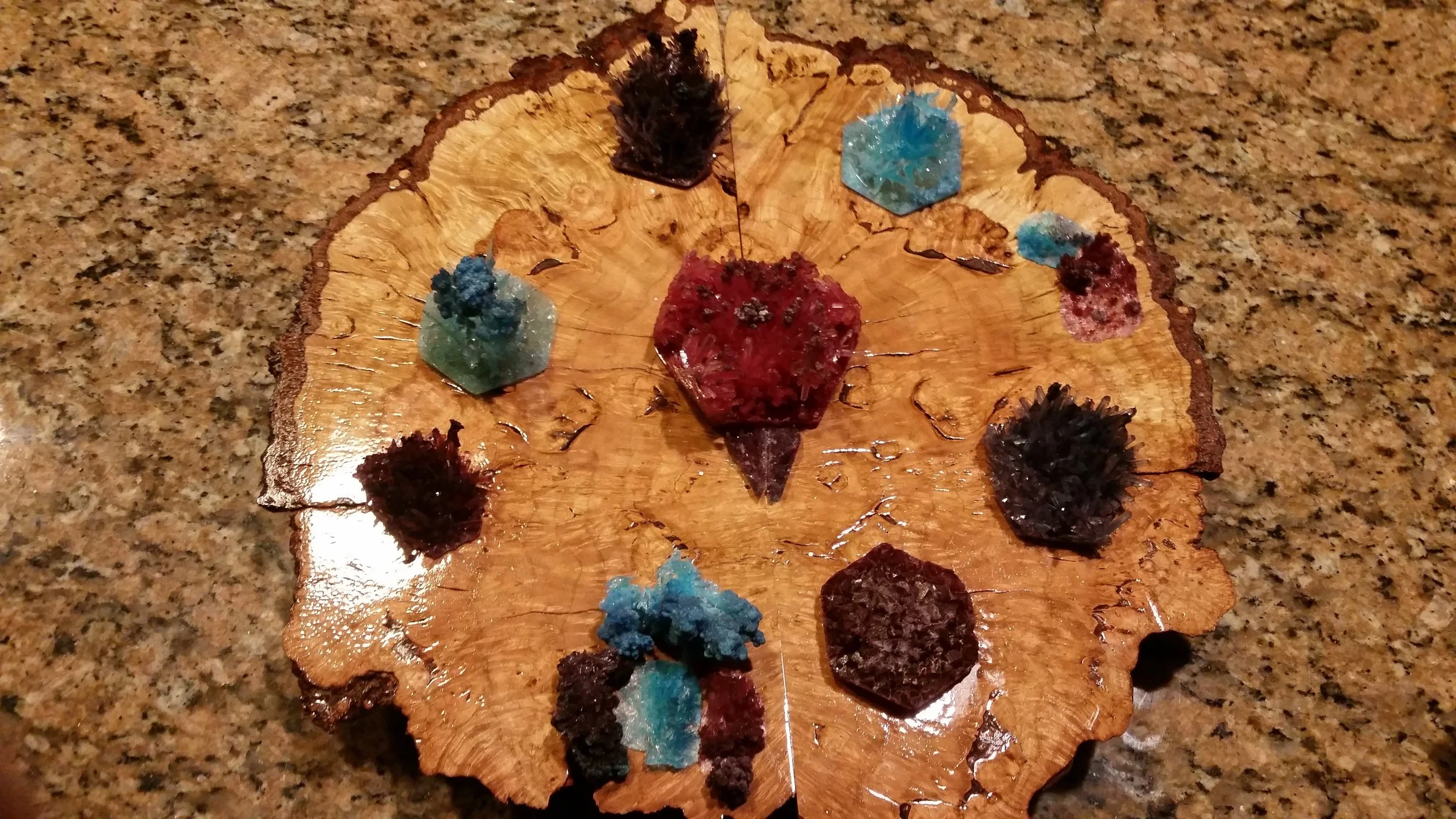A wooden tree cross-section with multiple clusters of colorful mineral or crystal specimens placed on it, arranged in a circular pattern.