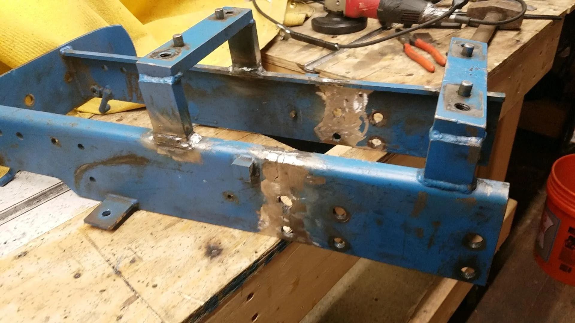 Welded blue metal frame on a workbench in a workshop, surrounded by tools, with visible weld marks and holes.