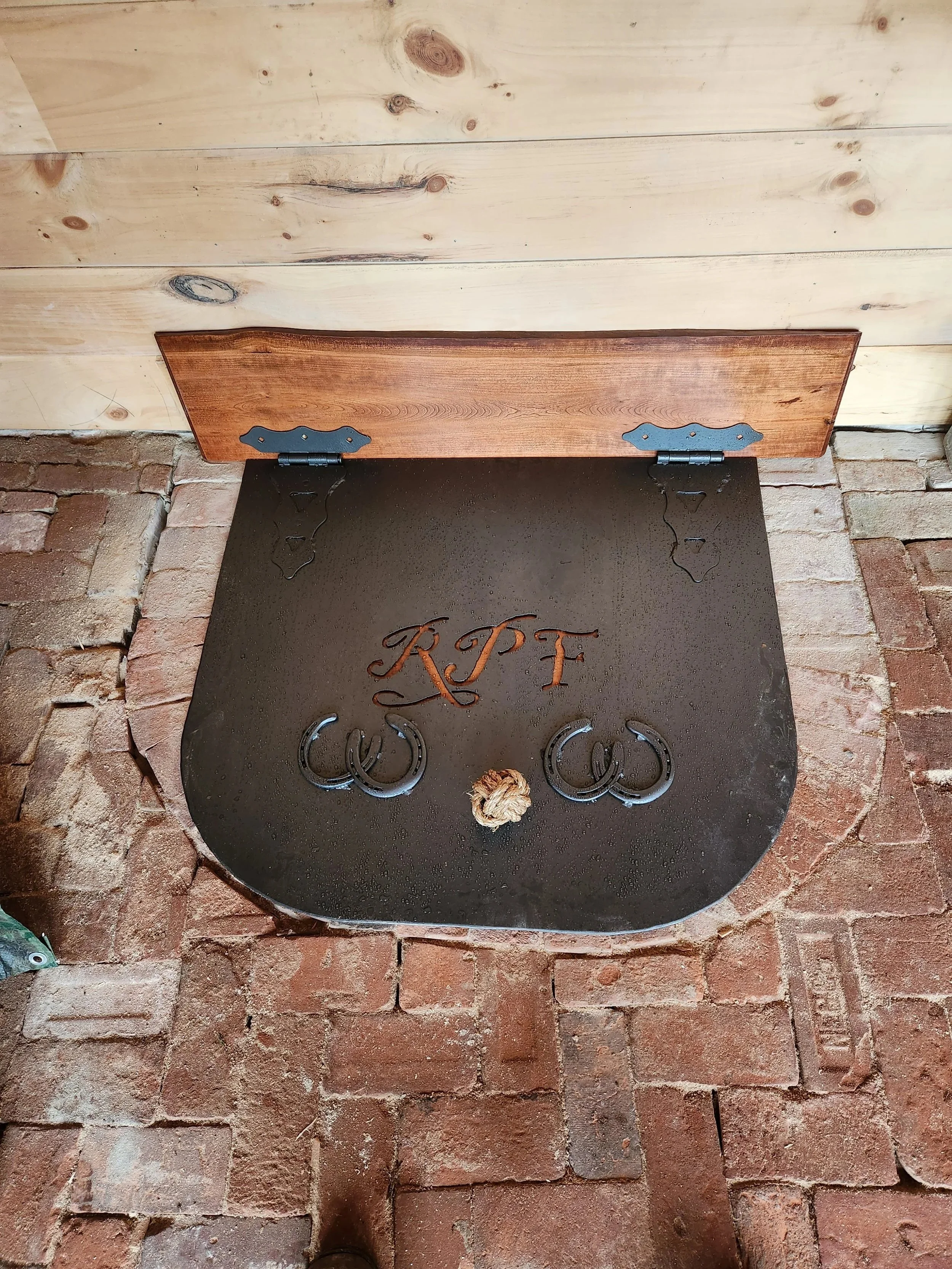 A black metal pet door with the initials 'KPF' and paw prints, mounted in a brick wall with a wooden door above.