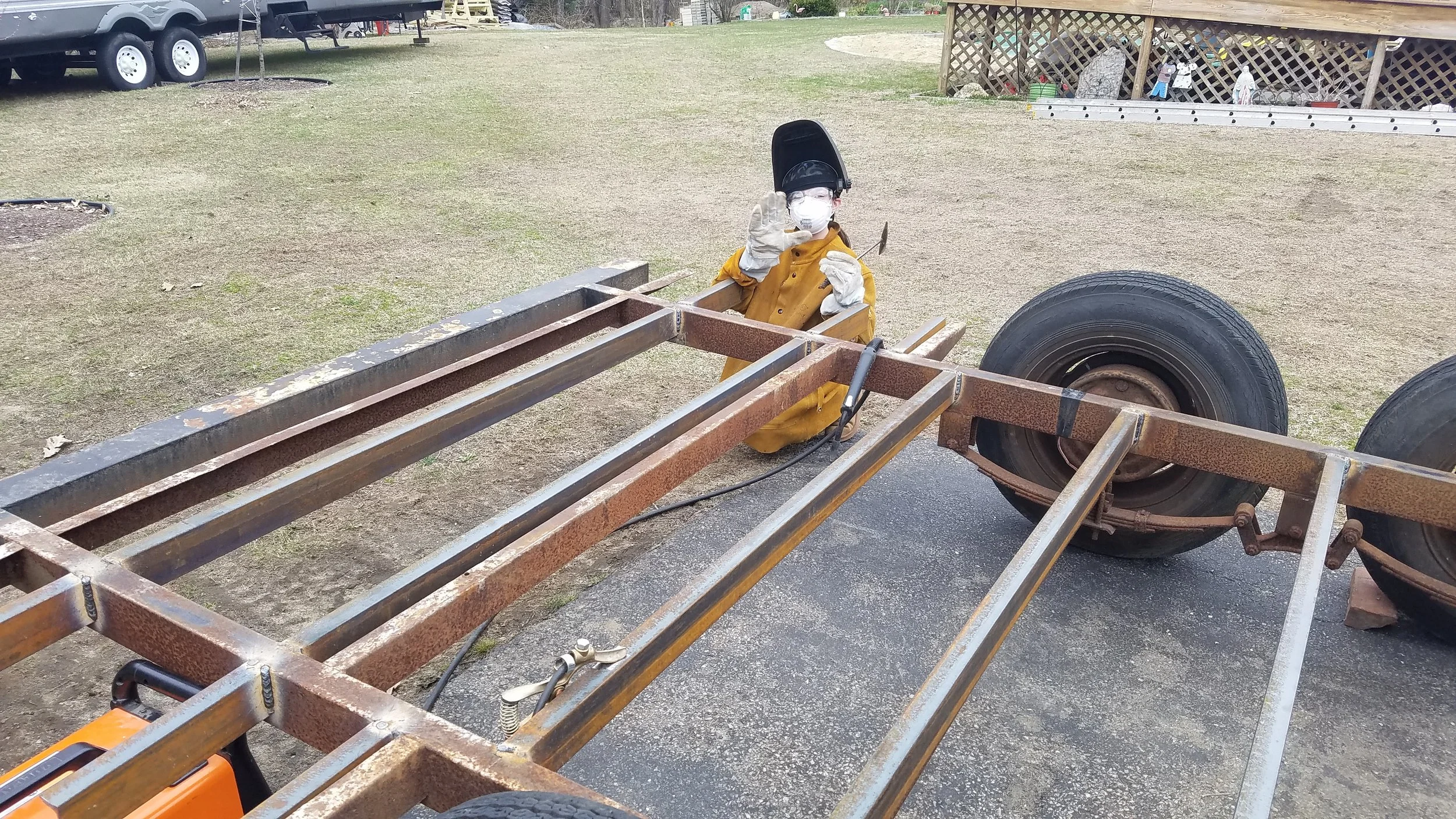 Person wearing protective gear welding on a rusty metal frame outdoors near large vehicle tires, with a yard and some structures in the background.