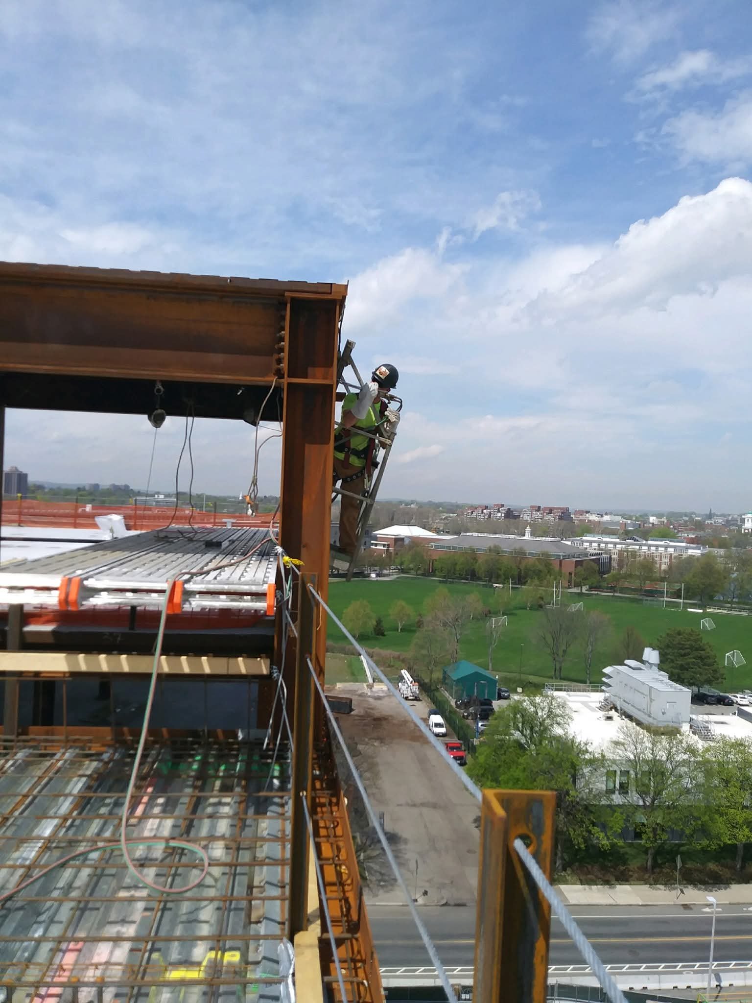 A construction worker wearing a helmet and harness is working on the exterior of a high-rise building, using a ladder to reach the upper levels.