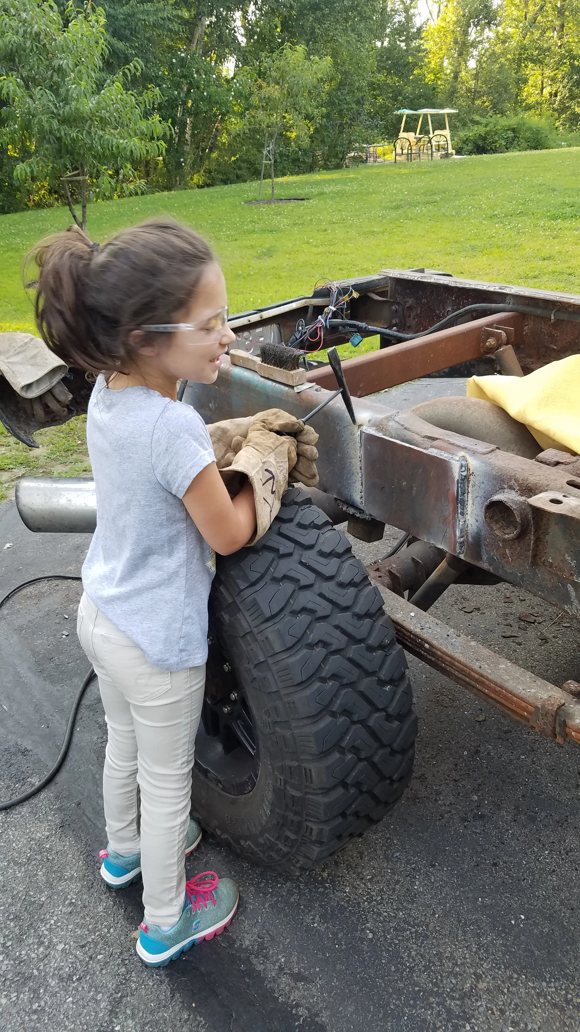 A smiling young girl wearing safety goggles and gloves is working on a rusty vehicle frame outdoors in a grassy area with trees in the background. She is holding a tool near the vehicle's large tire.