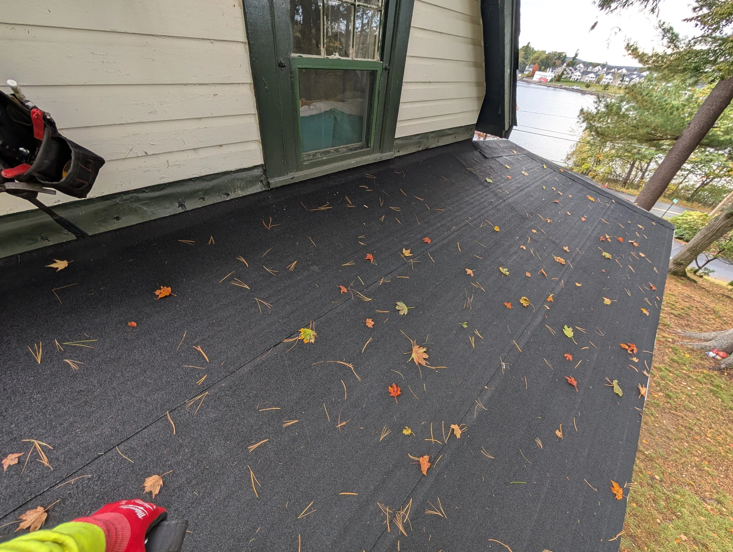 Black rubber roofing on a house's flat roof, scattered with fallen autumn leaves, with a view of trees, a river, and houses in the background.