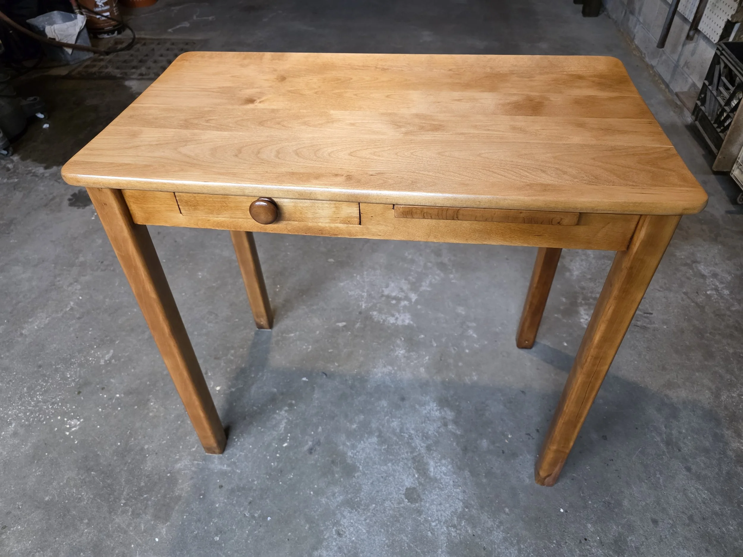 A wooden table with a single drawer and a round knob, standing on a concrete floor.