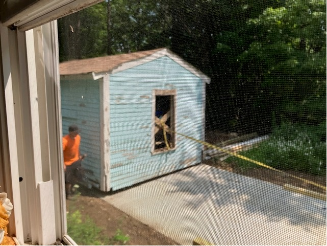 View through a window screen showing a small blue shed being built with a person in an orange shirt nearby.