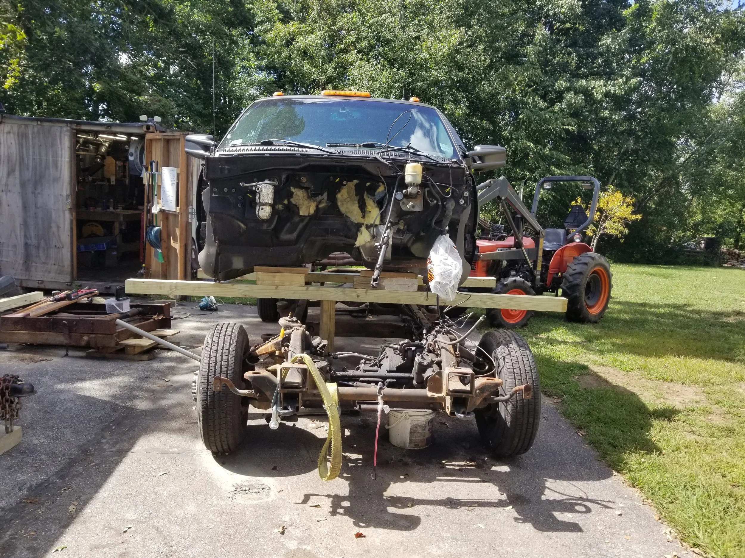 A vehicle is being disassembled or repaired outdoors, with the front wheels and engine removed. The vehicle's cabin is elevated on a wooden framework next to a tractor on a grassy area.