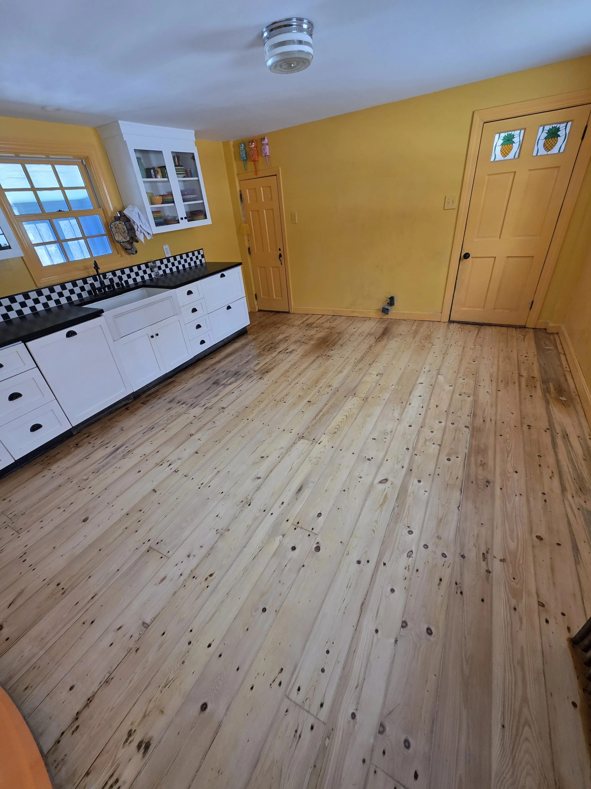Empty kitchen with natural wood flooring, yellow walls, white cabinet with black countertop and checkered backsplash, and two stained glass pineapple window panels on a wooden door.