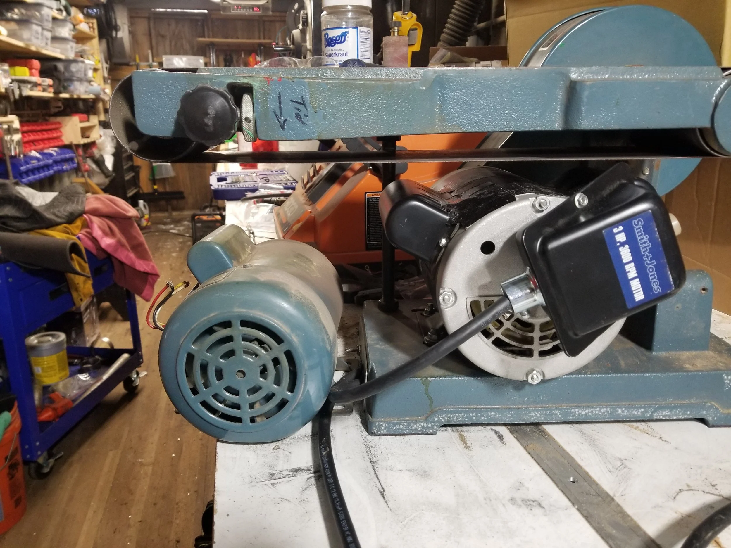 Close-up of a blue metal band saw with a motor, placed on a workbench in a workshop, with tools and supplies in the background.