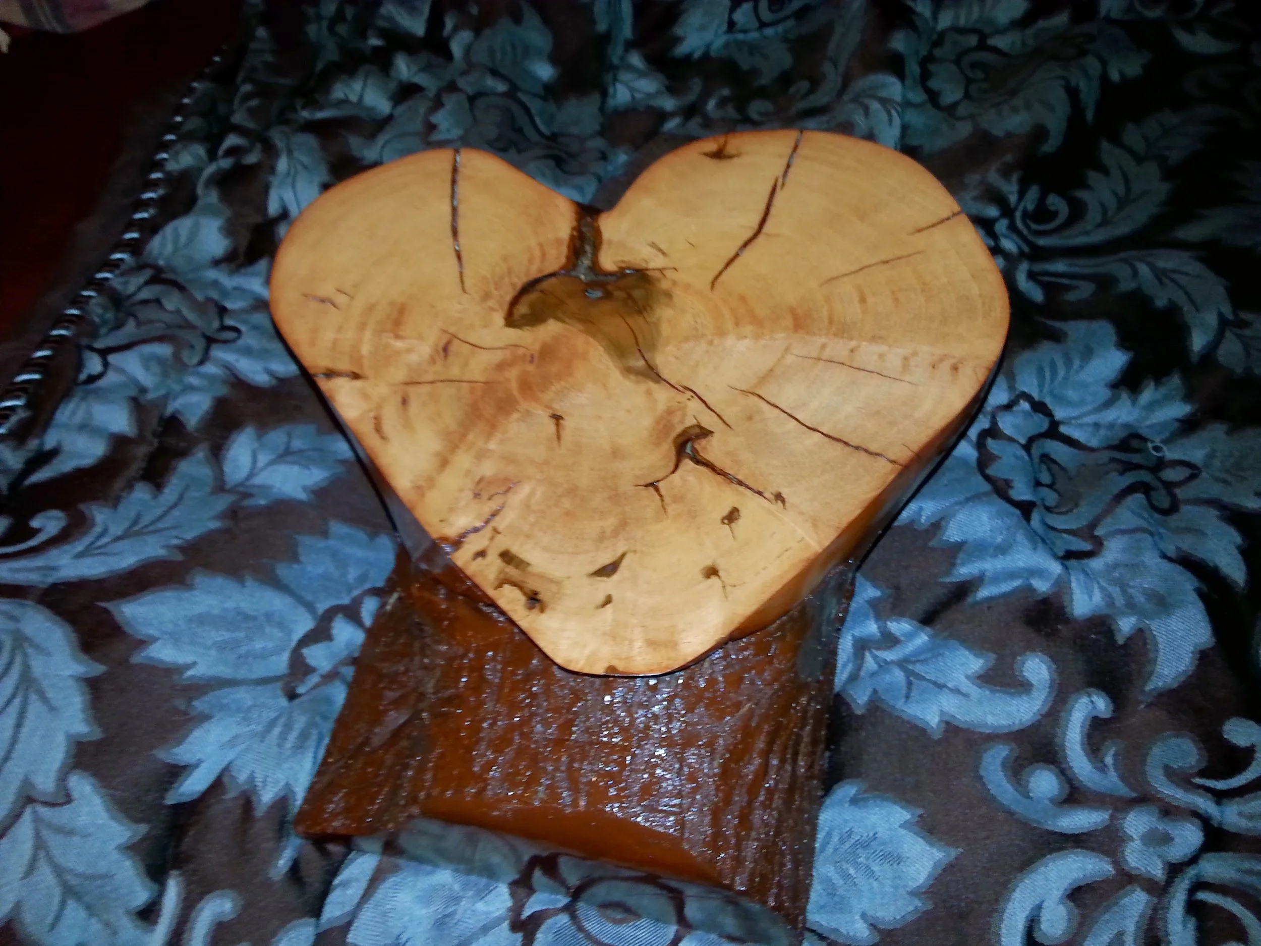 A heart-shaped piece of wood with visible growth rings and cracks, placed on a dark floral fabric surface.