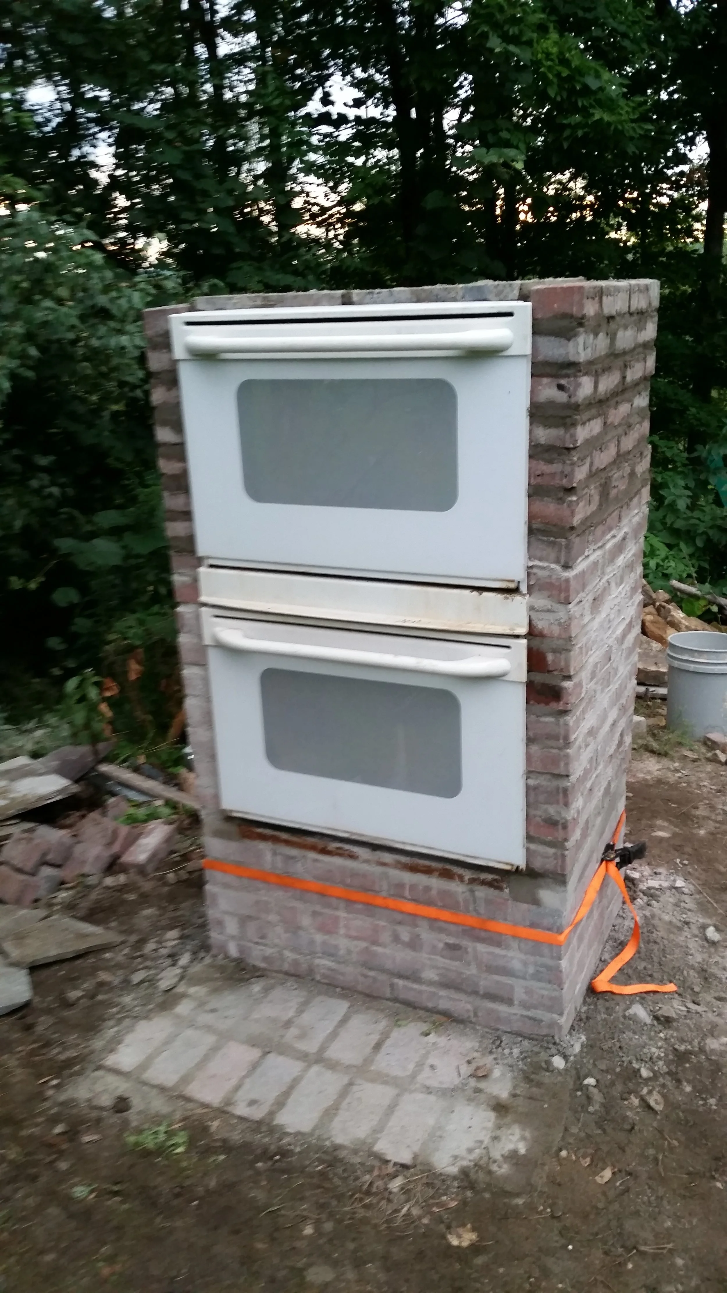 Outdoor brick structure with two white oven doors, surrounded by trees and construction materials.
