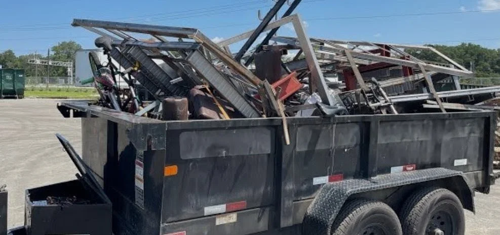 A black trailer filled with discarded metal and scrap materials parked on a paved lot under a clear blue sky.