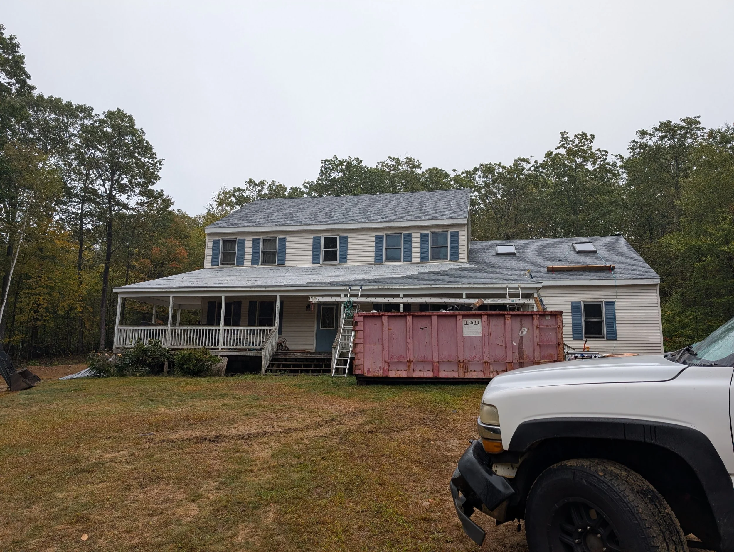House under renovation with a large red dumpster in front, construction ladder and roof work in progress, surrounded by trees and an overcast sky.