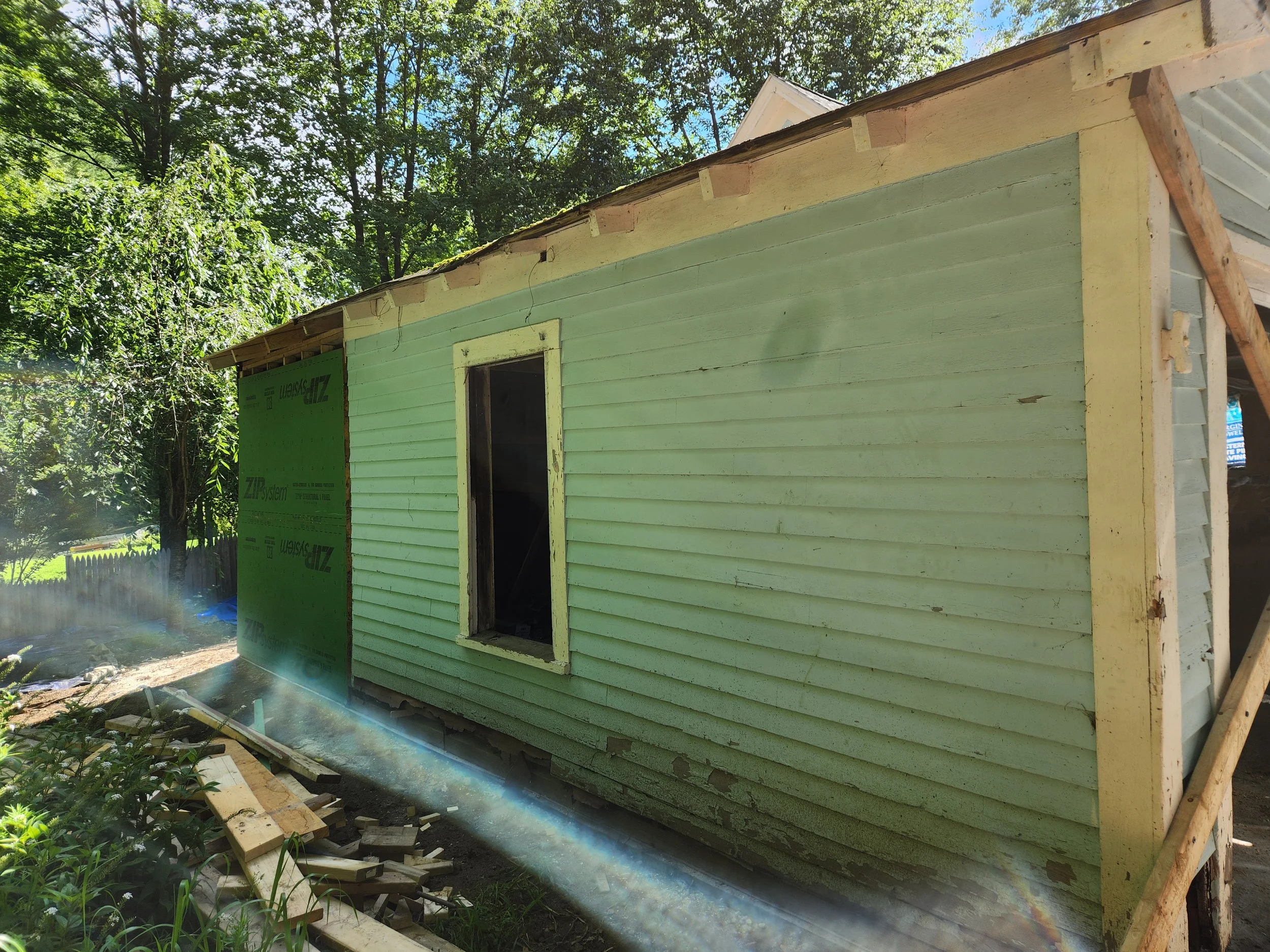 A house under construction with green siding, a window frame without glass, surrounded by trees and construction materials on the ground.