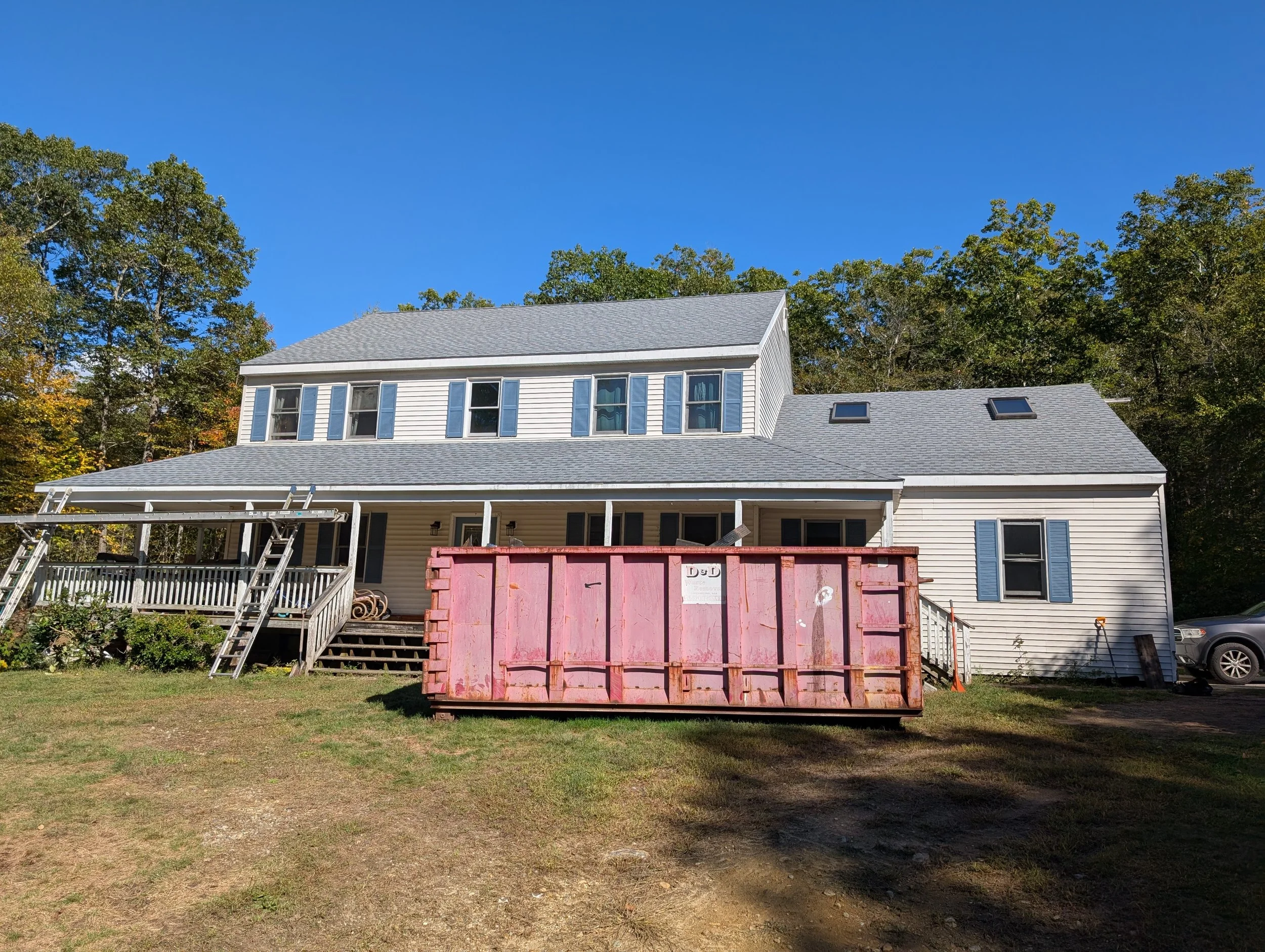 A large house under renovation with a pink dumpster in front, surrounded by trees on a clear day.