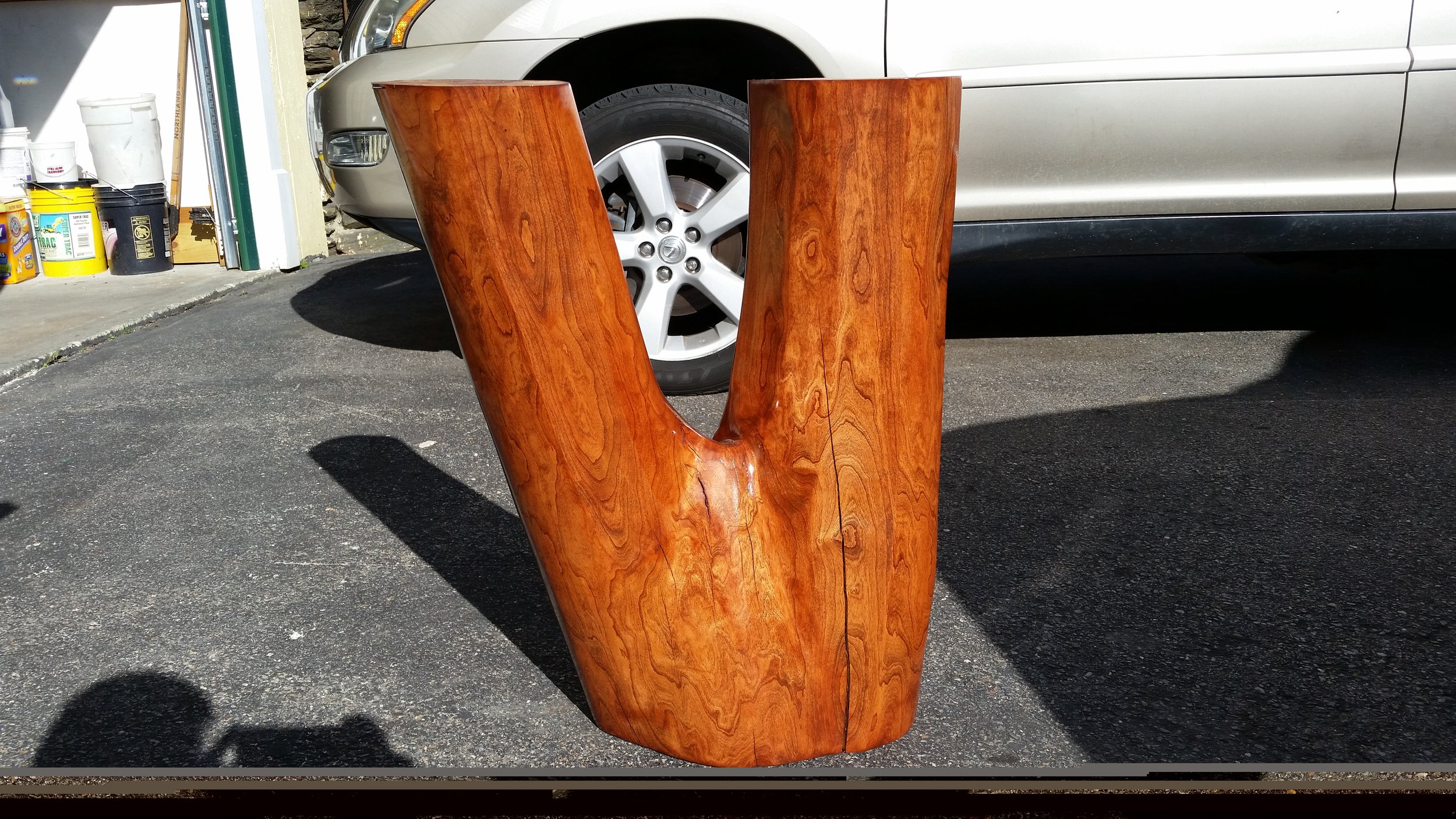 A large wooden V-shaped sculpture with a carved hole at the top, placed on an asphalt driveway, with a silver car and storage containers in the background.