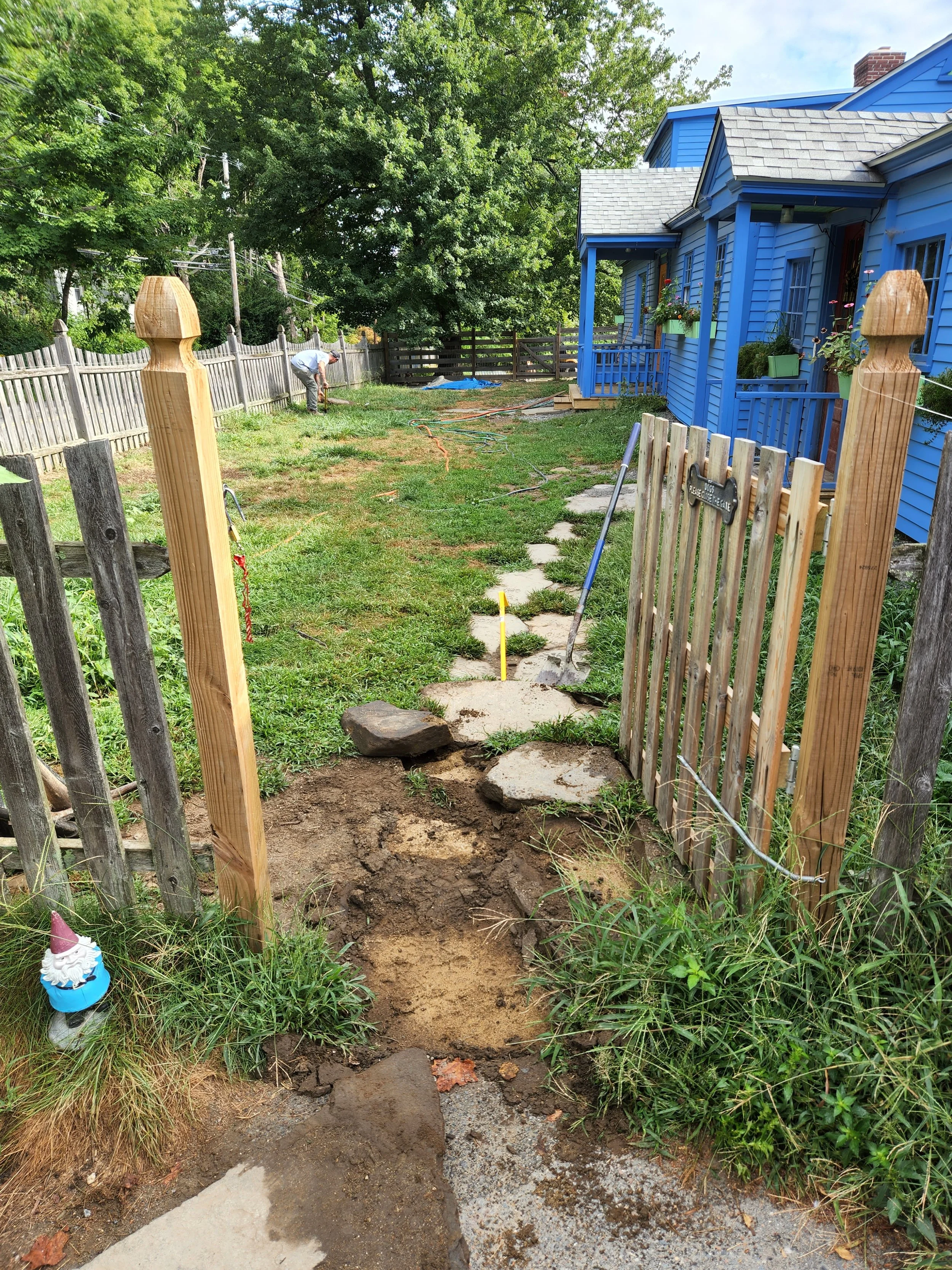 Person working on outdoor stone pathway in yard next to a blue house, with construction tools and materials around, fenced yard with trees in background.