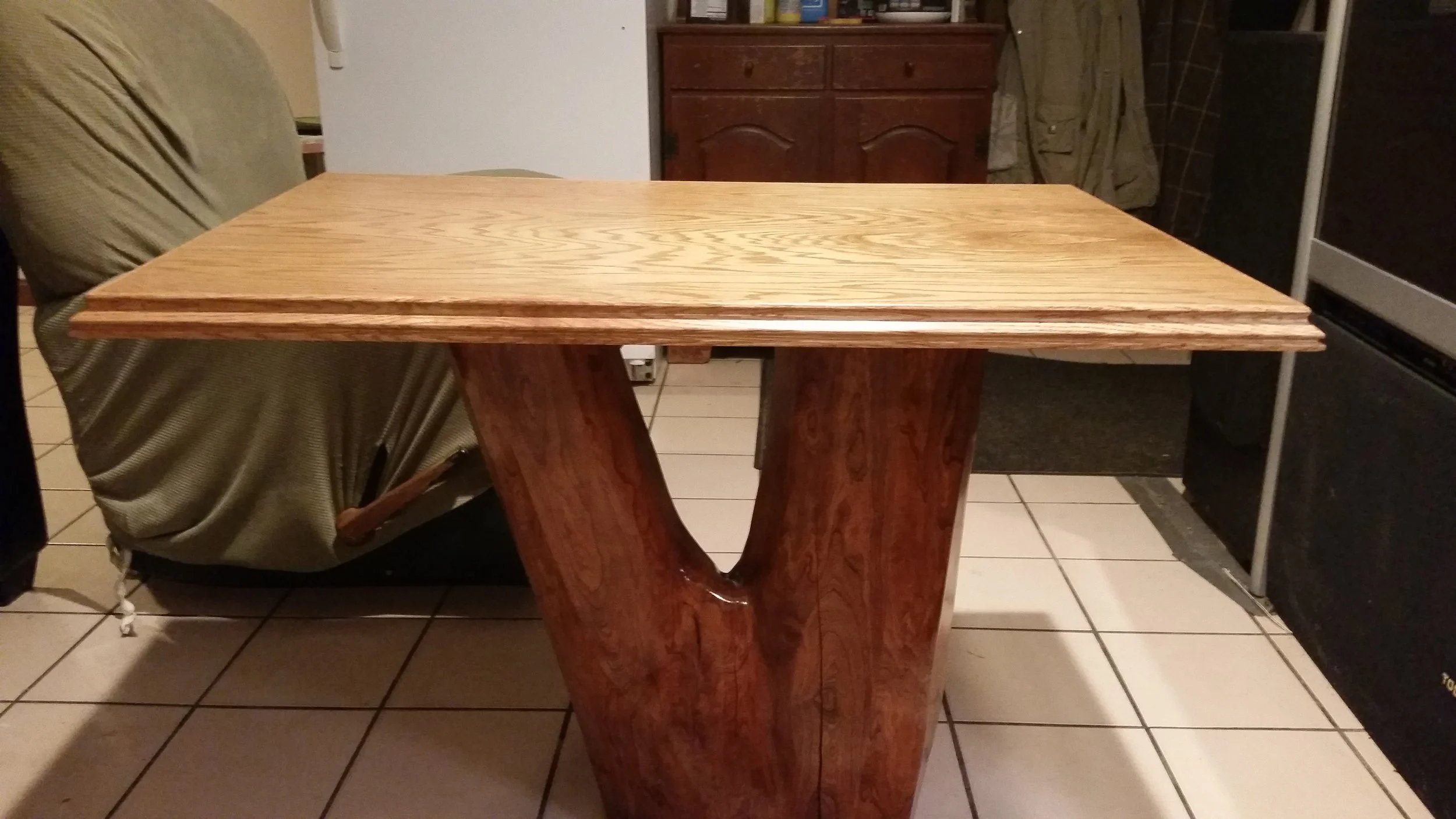 A wooden table with a thick, flat top and a unique V-shaped pedestal base, situated in a kitchen with tiled floor, a couch, and kitchen cabinets.