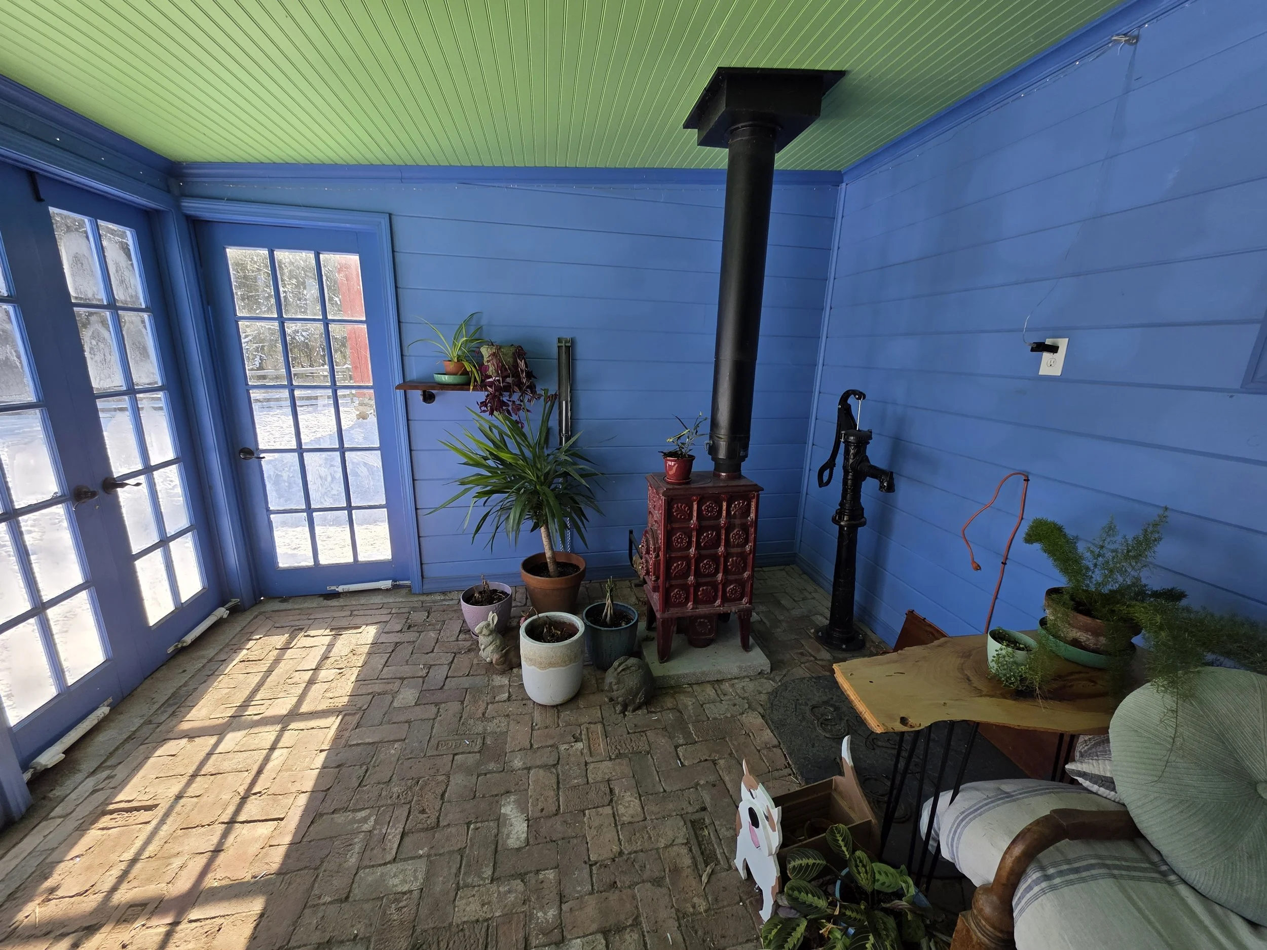 Sunlit porch with blue walls and brick floor, decorated with potted plants, a wood stove, and a wooden armchair with a pillow.