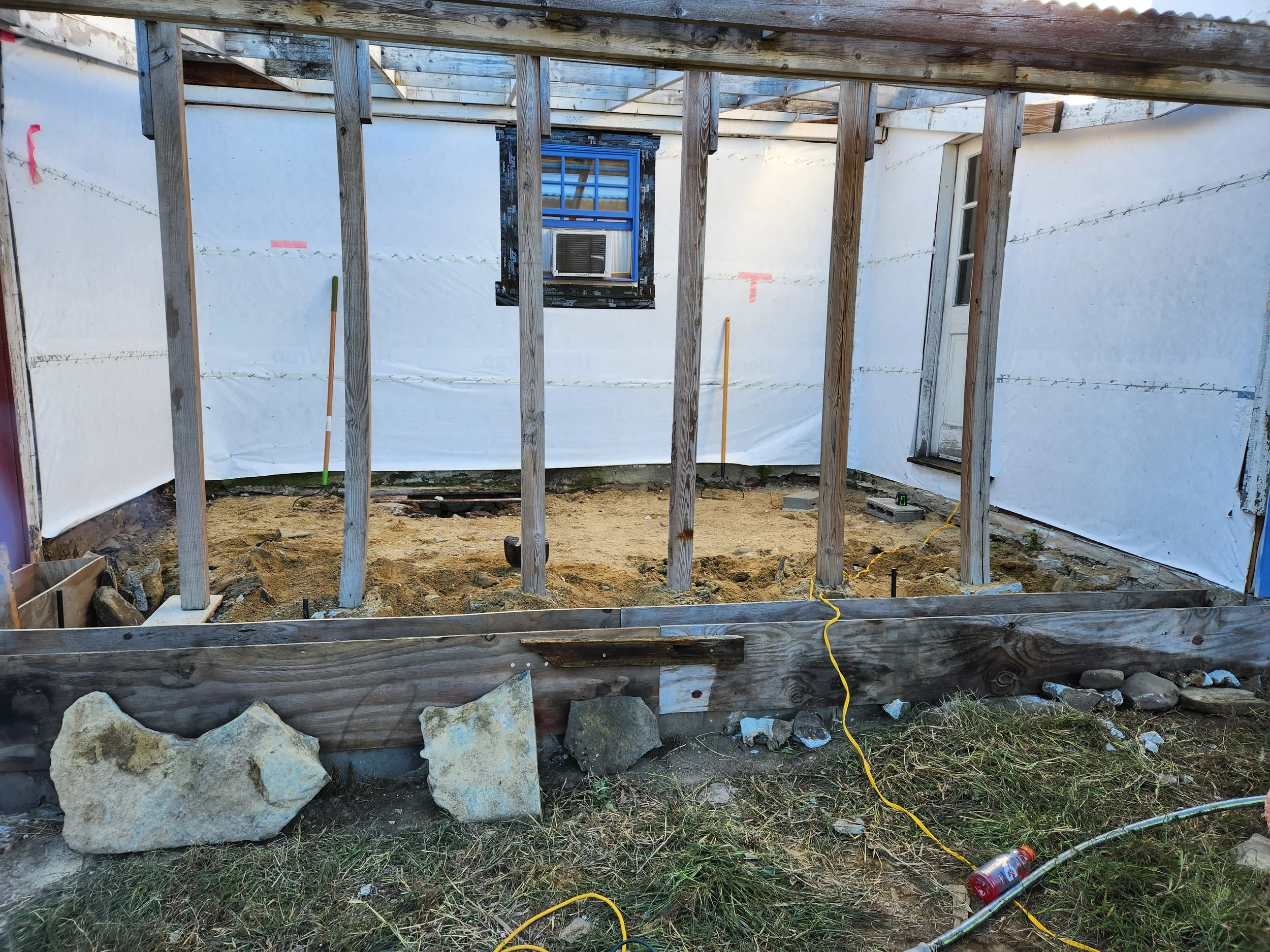 Construction site with a wooden frame, sand on the ground, and a window with an air conditioning unit, wrapped with plastic sheeting.