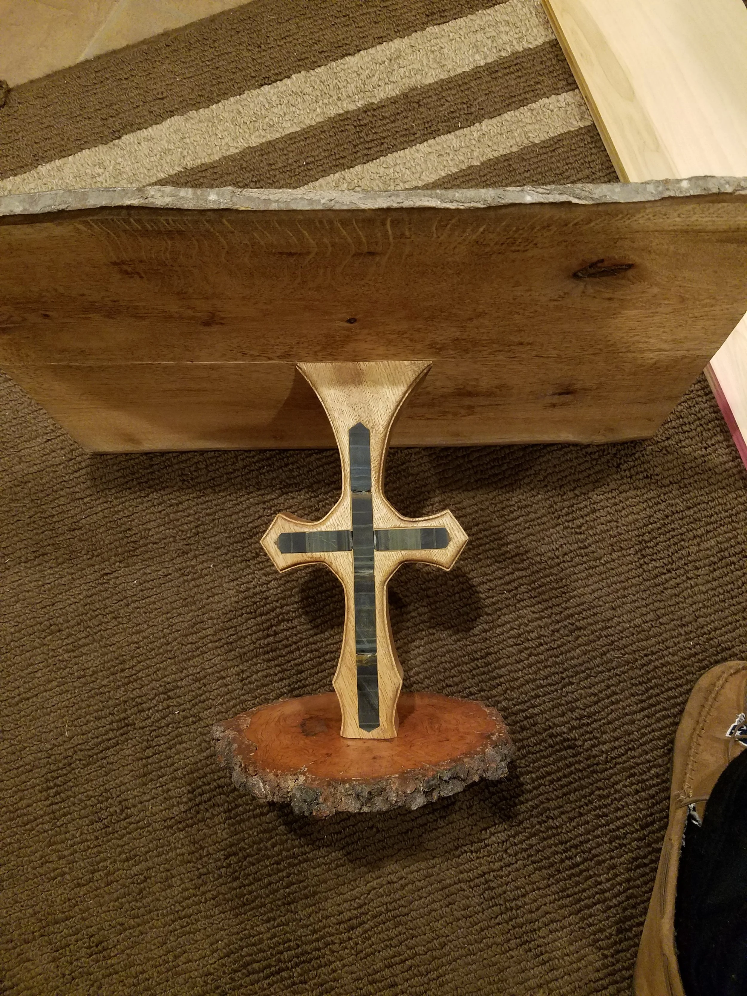 A decorative wooden cross with black inlay inlaid in the wood, mounted on a carved wooden base, placed on a brown textured carpet.