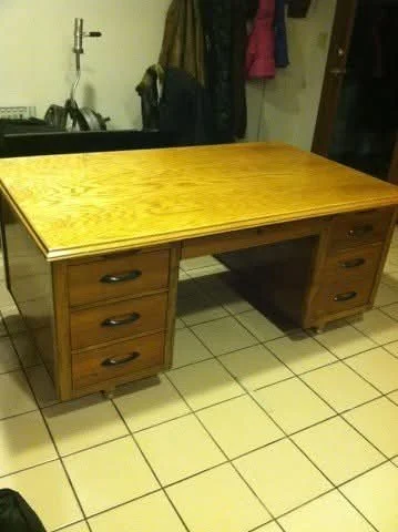 Wooden desk with a light-colored top and multiple drawers on both sides, sitting on a tiled floor in a room with coat hooks and clothing in the background.