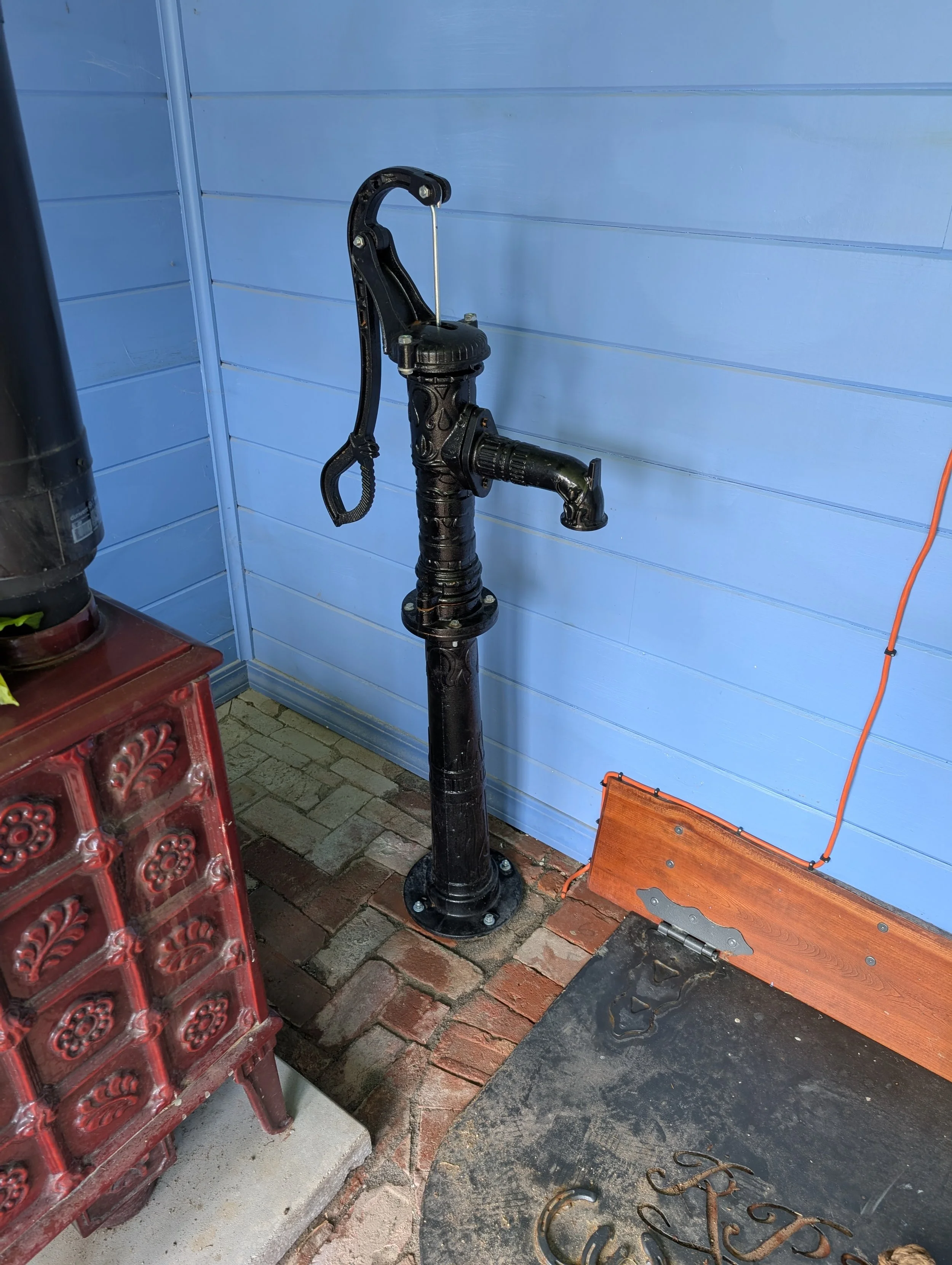 Decorative black water fountain with a spout, situated next to a blue wall on a brick and concrete floor, with a red decorative piece and a black mat nearby.