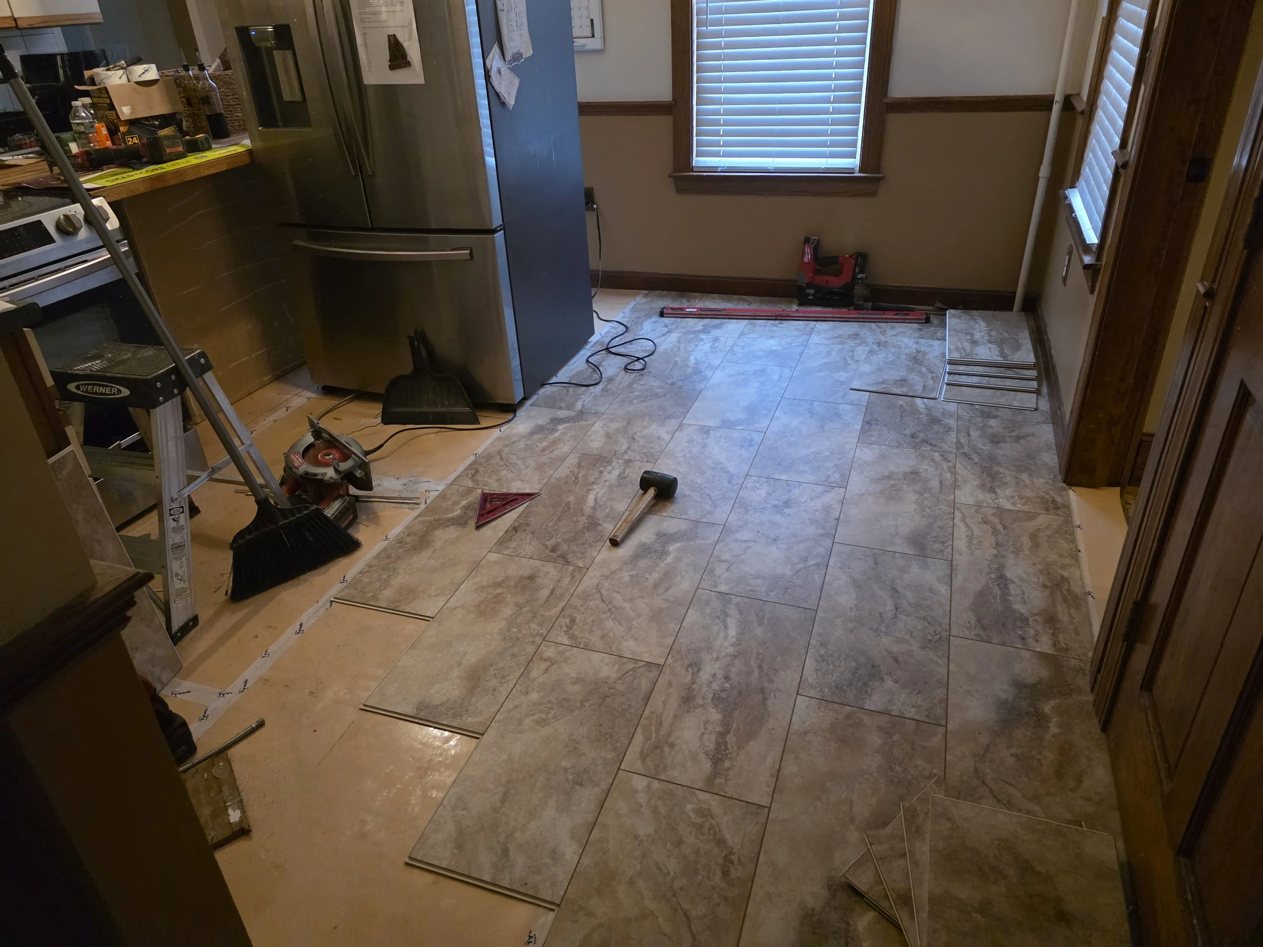 A kitchen with tile flooring installation in progress, tools including a hammer, leveling tool, broom, and a saw scattered on the floor, and various kitchen appliances and cabinets present.
