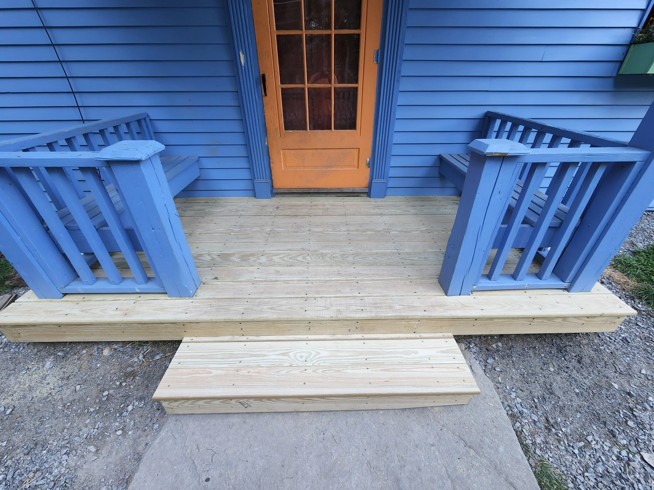 Newly built wooden porch with blue railings and a small step leading to a door painted in a tan color, attached to a blue house with horizontal panel siding.