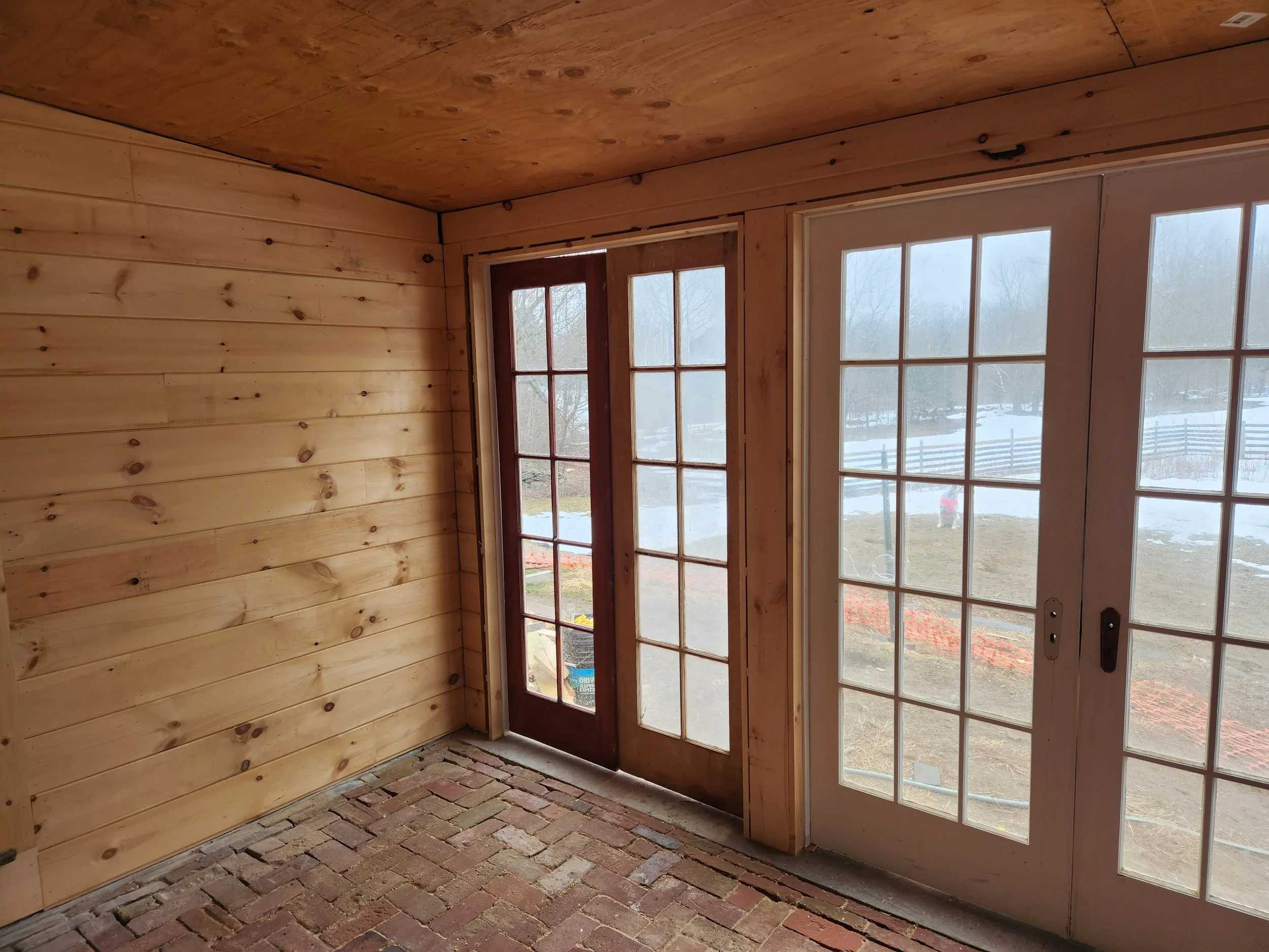 Interior view of a room with wooden walls and ceiling, brick floor, and French doors leading outside; some snow and fencing visible through the glass.