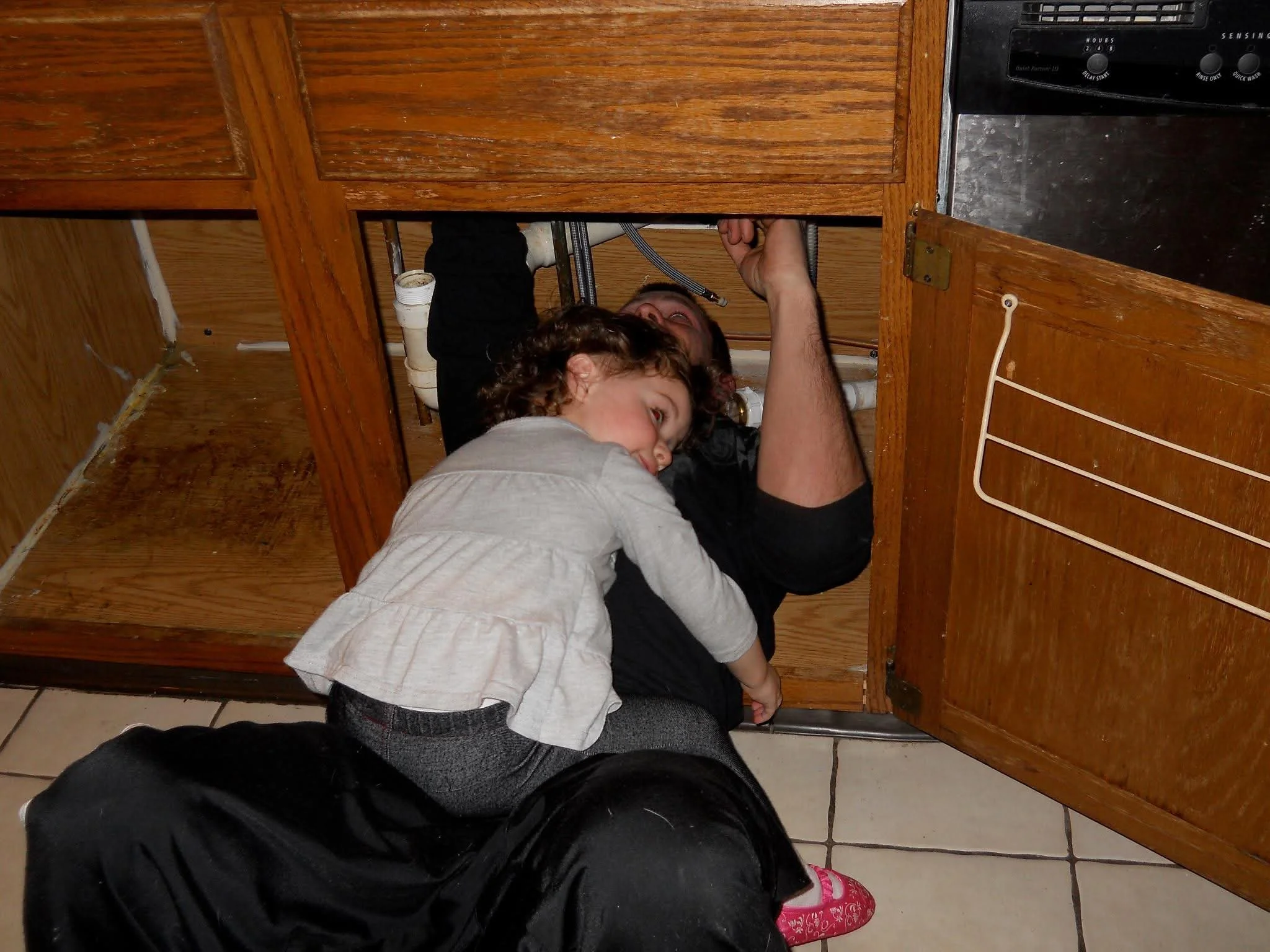 A man and a young girl fixing or inspecting pipes under a kitchen sink.