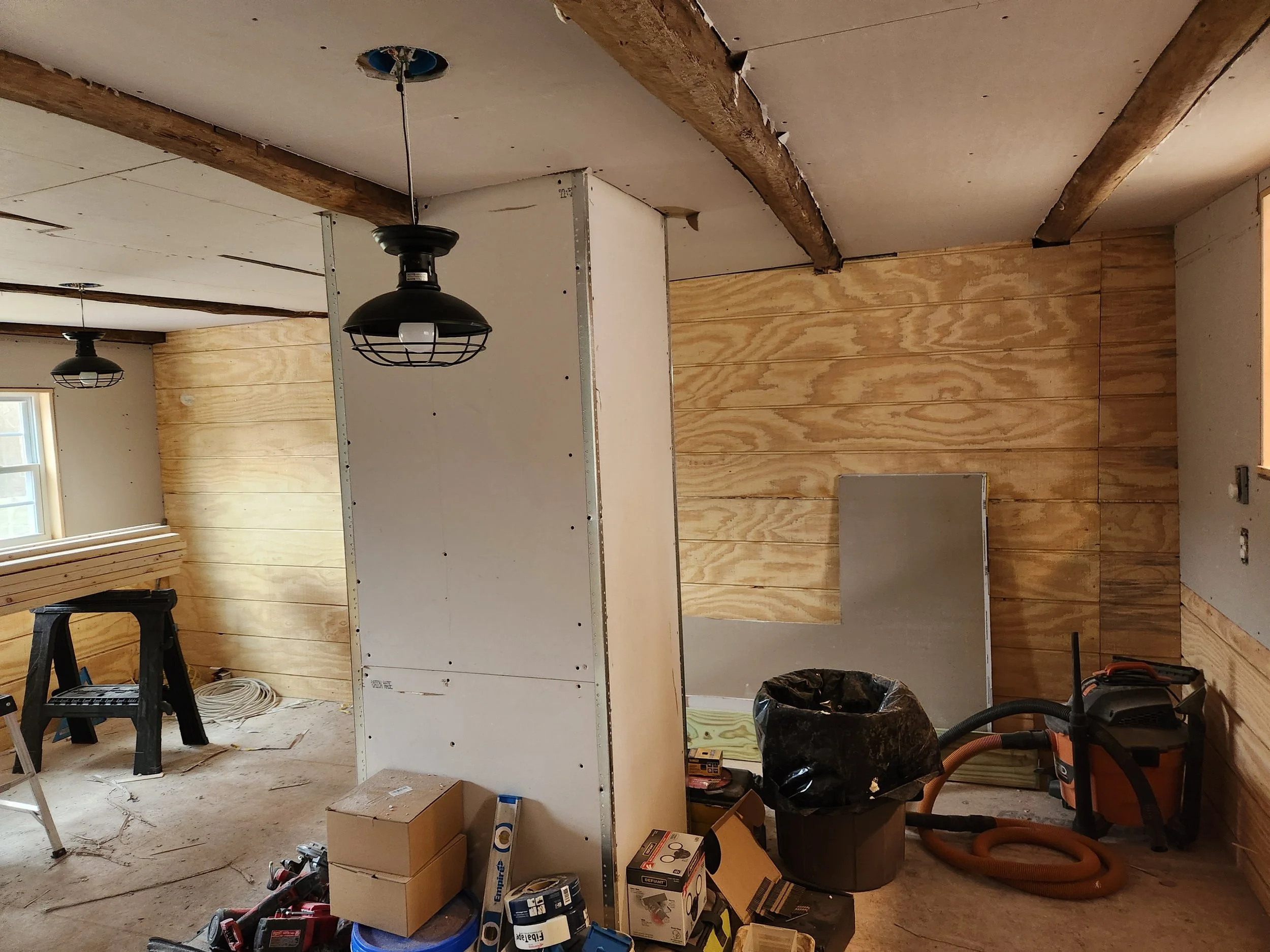 Interior of a room under construction with wooden wall paneling, unfinished ceiling, construction tools, boxes, and equipment scattered around.