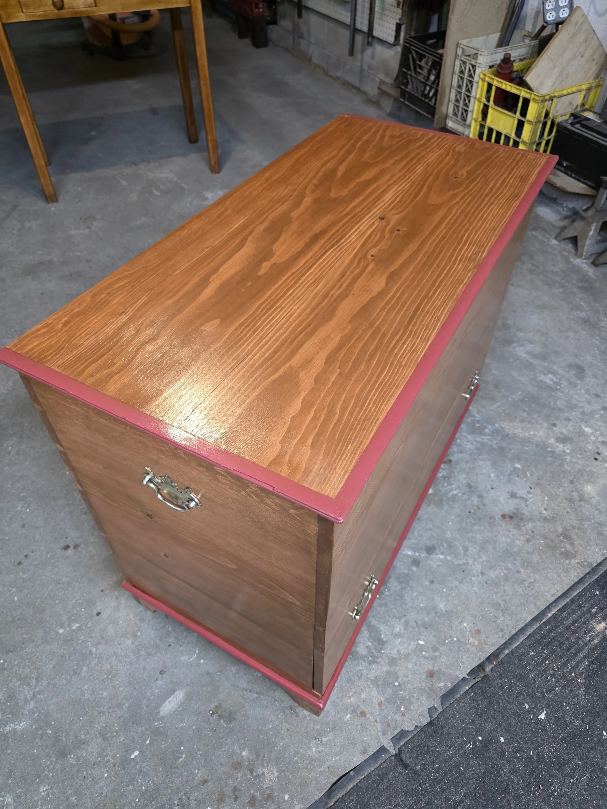Wooden storage cabinet with brass handles, placed on a concrete garage floor, with a red trim along the top edge and bottom base, in a garage or workshop setting.