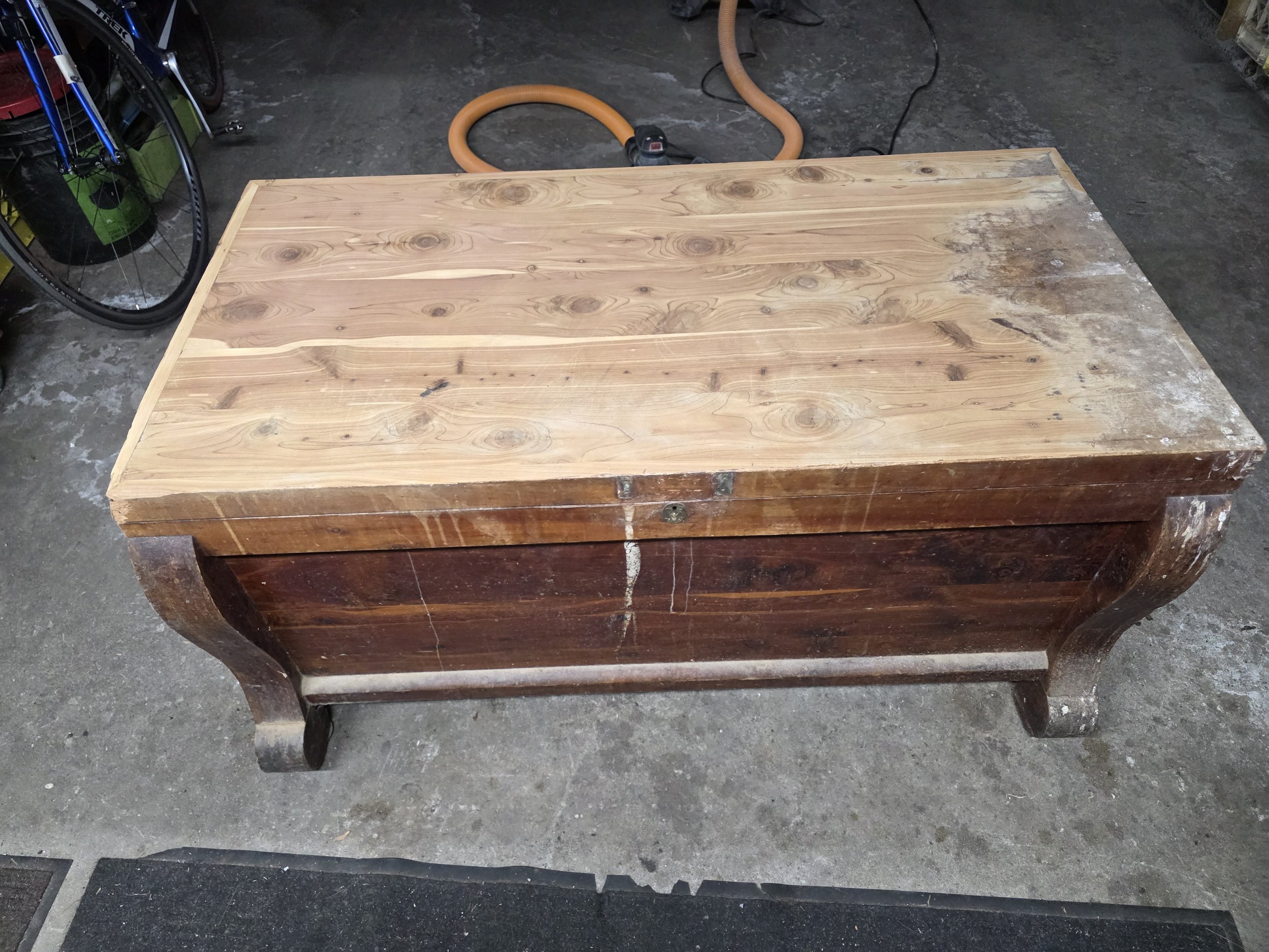 A wooden chest or storage box with a flat top, standing on curved legs in a workshop. The surface shows some stains and marks.