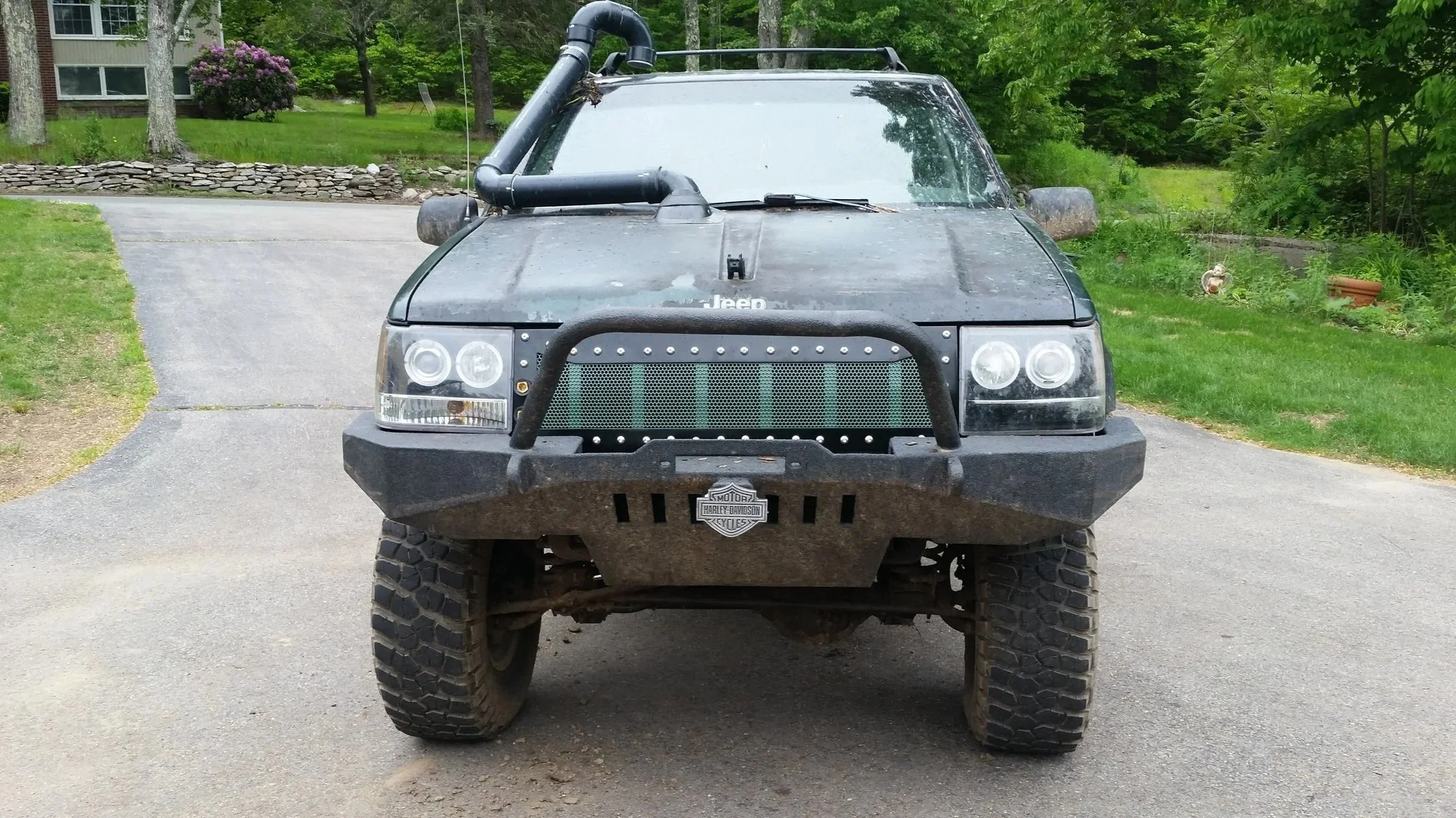 Front view of a black Jeep with off-road modifications, including a heavy-duty bumper, a snorkel, large tires, and a mesh grille, parked on a driveway with greenery in the background.