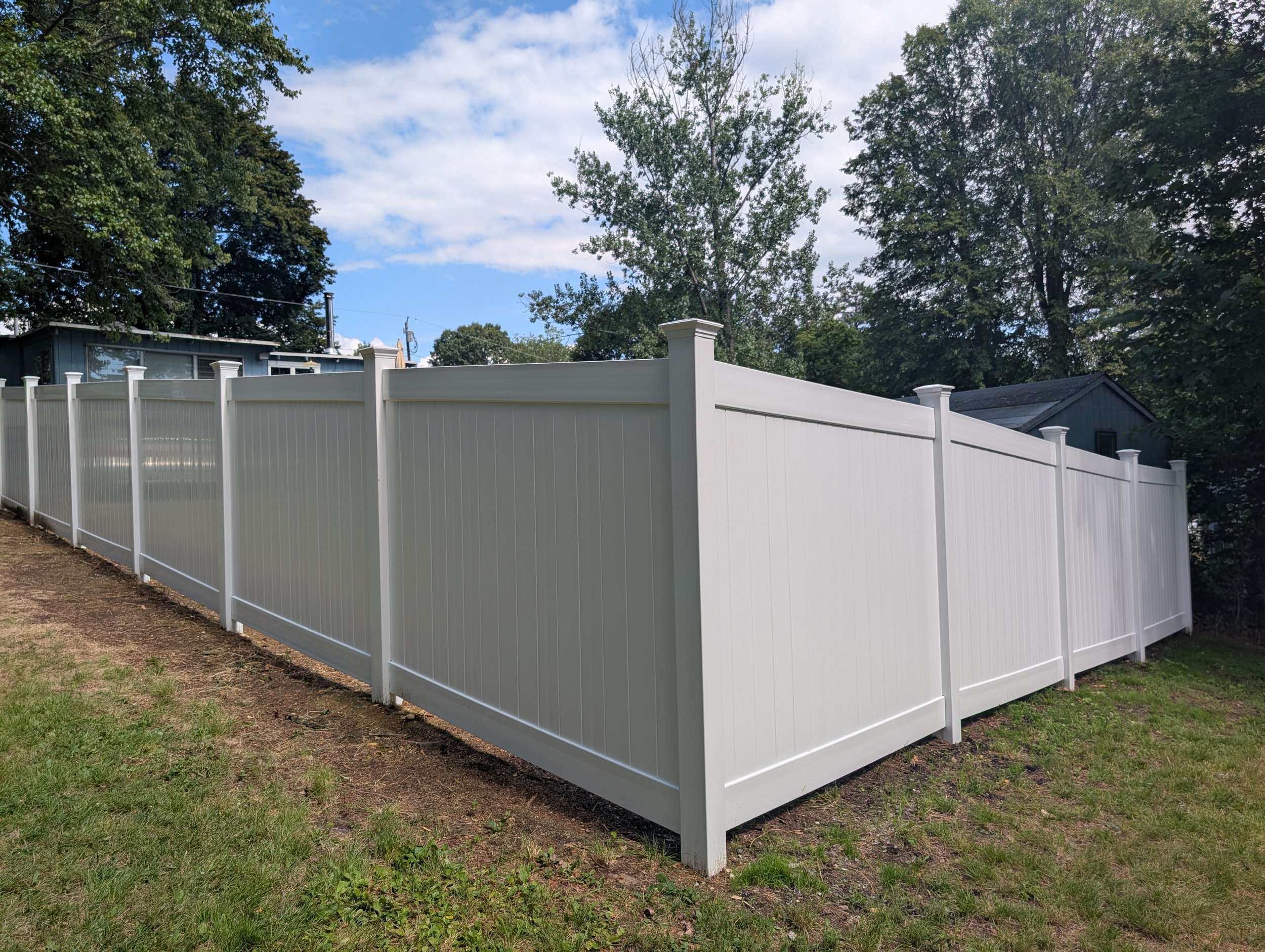 A white privacy fence surrounds a backyard with grass and a house in the background, under a partly cloudy sky.