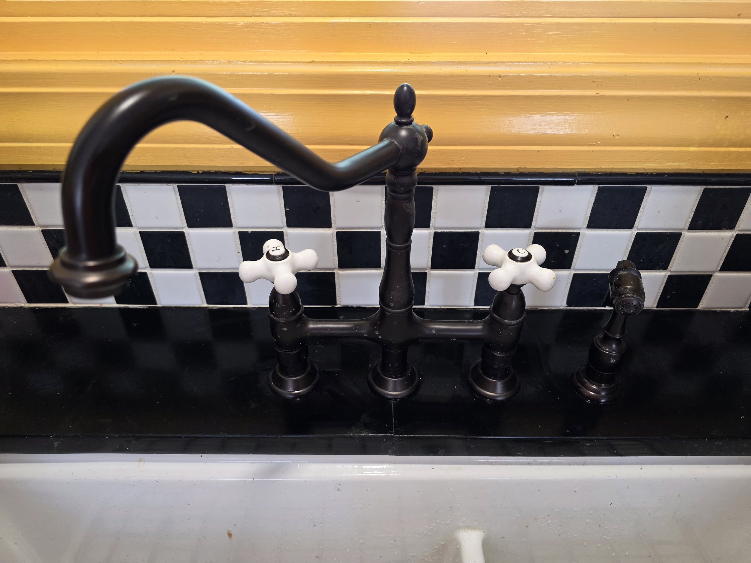 Black kitchen sink faucet with two white handles, mounted on black countertop with checkered black and white tile backsplash and wooden wall above.